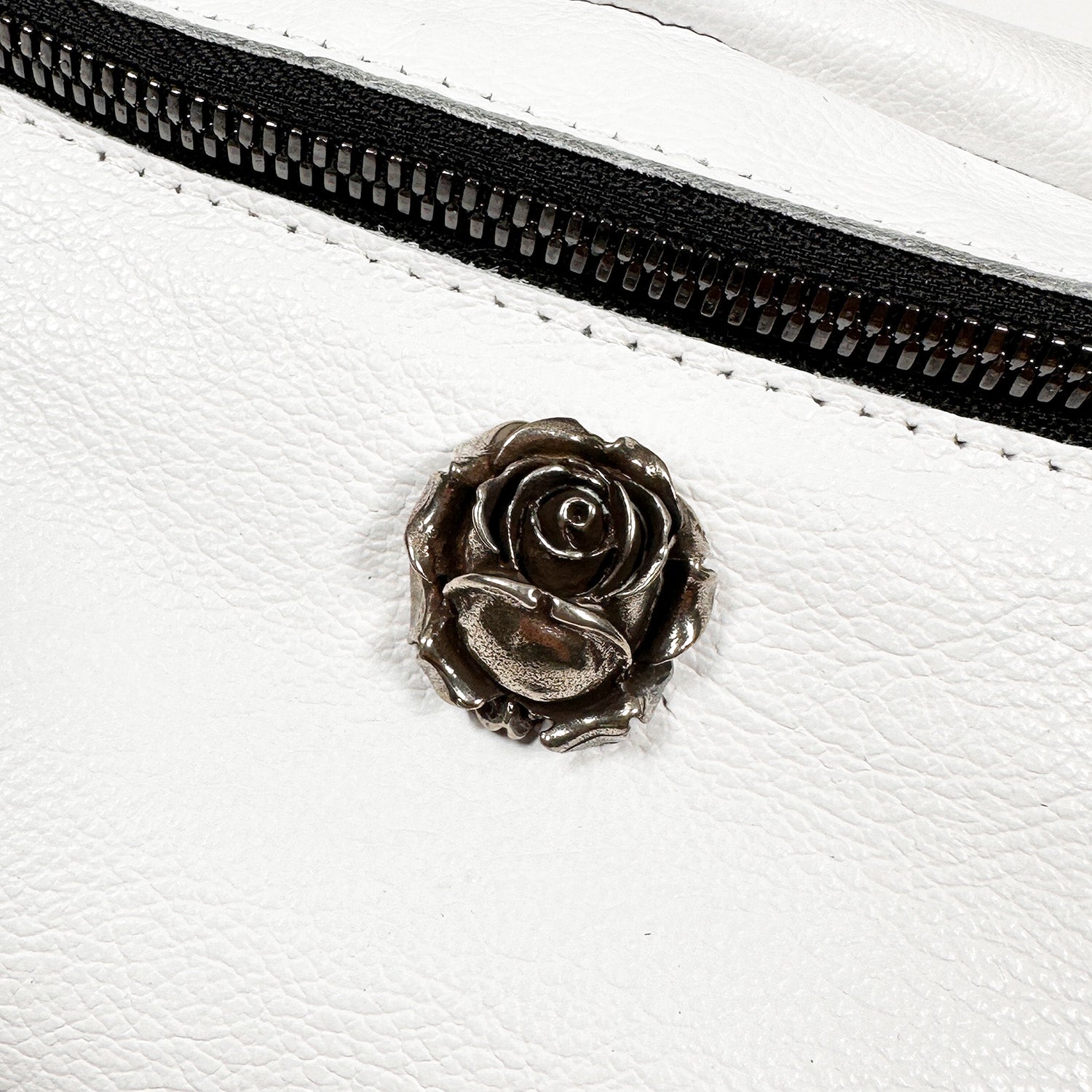 The "STELLA - white - rose" by evokewomen features an Italian leather surface in white, complemented by a black zipper, a metallic rose-shaped button, and an adjustable strap.