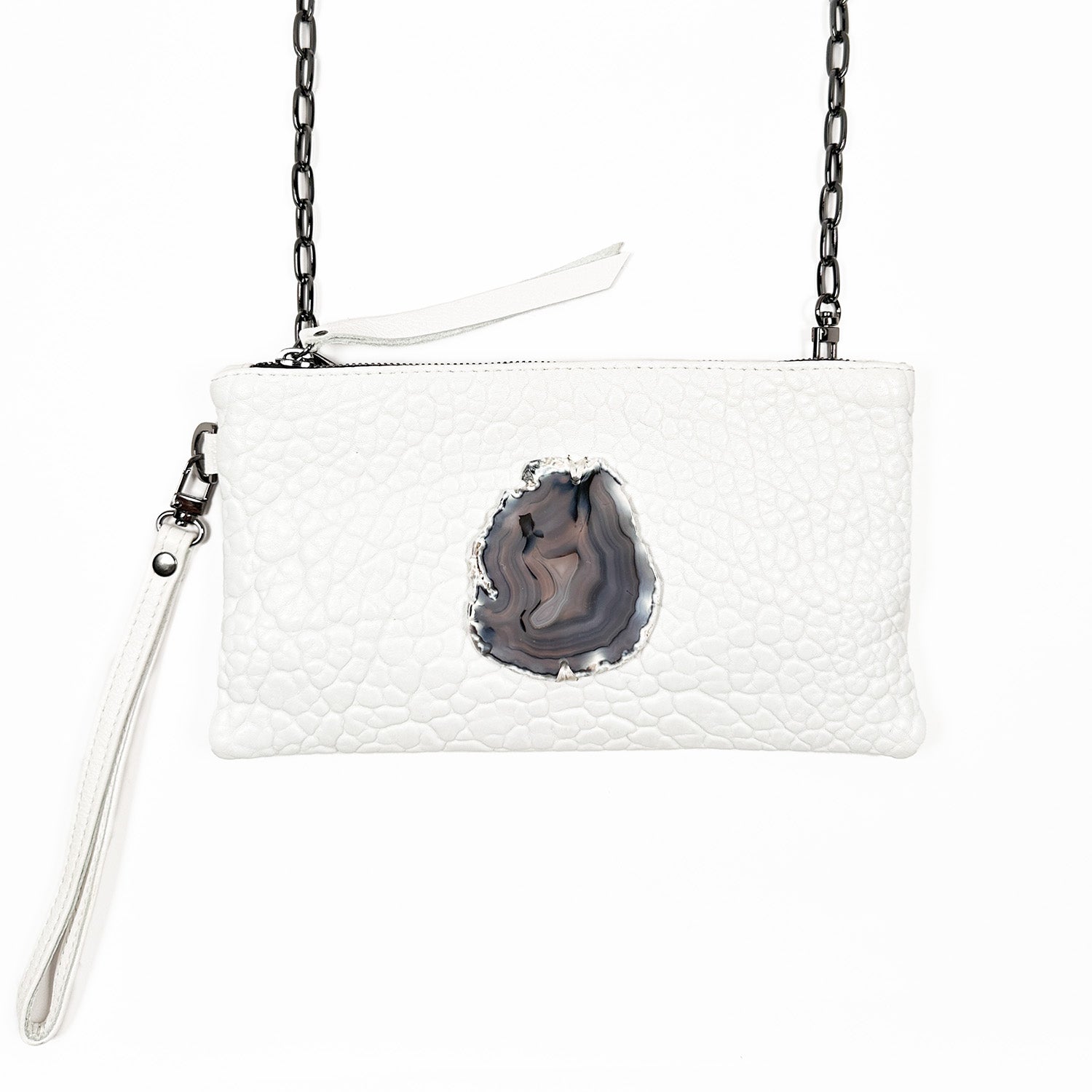The VICTORIA - white by evokewomen is a stylish textured clutch adorned with a dark, irregular agate stone accent at its center. Its versatile design includes a chain strap and wristlet, allowing it to effortlessly transform into a chic crossbody bag suitable for any occasion.