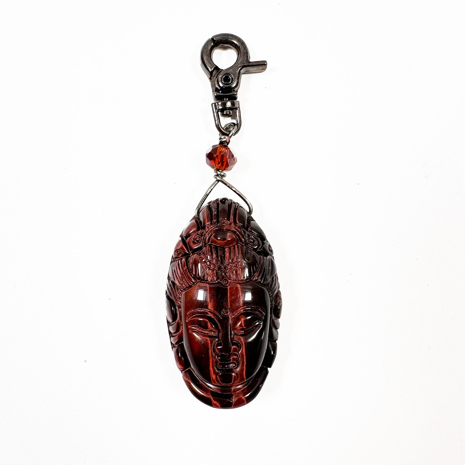 The Buddha Head Charm by evokewomen is a brown mask keychain featuring a carved face and adorned with a Red Tigereye bead. It includes a metal clasp, making it an easy clip-on charm against a white background.