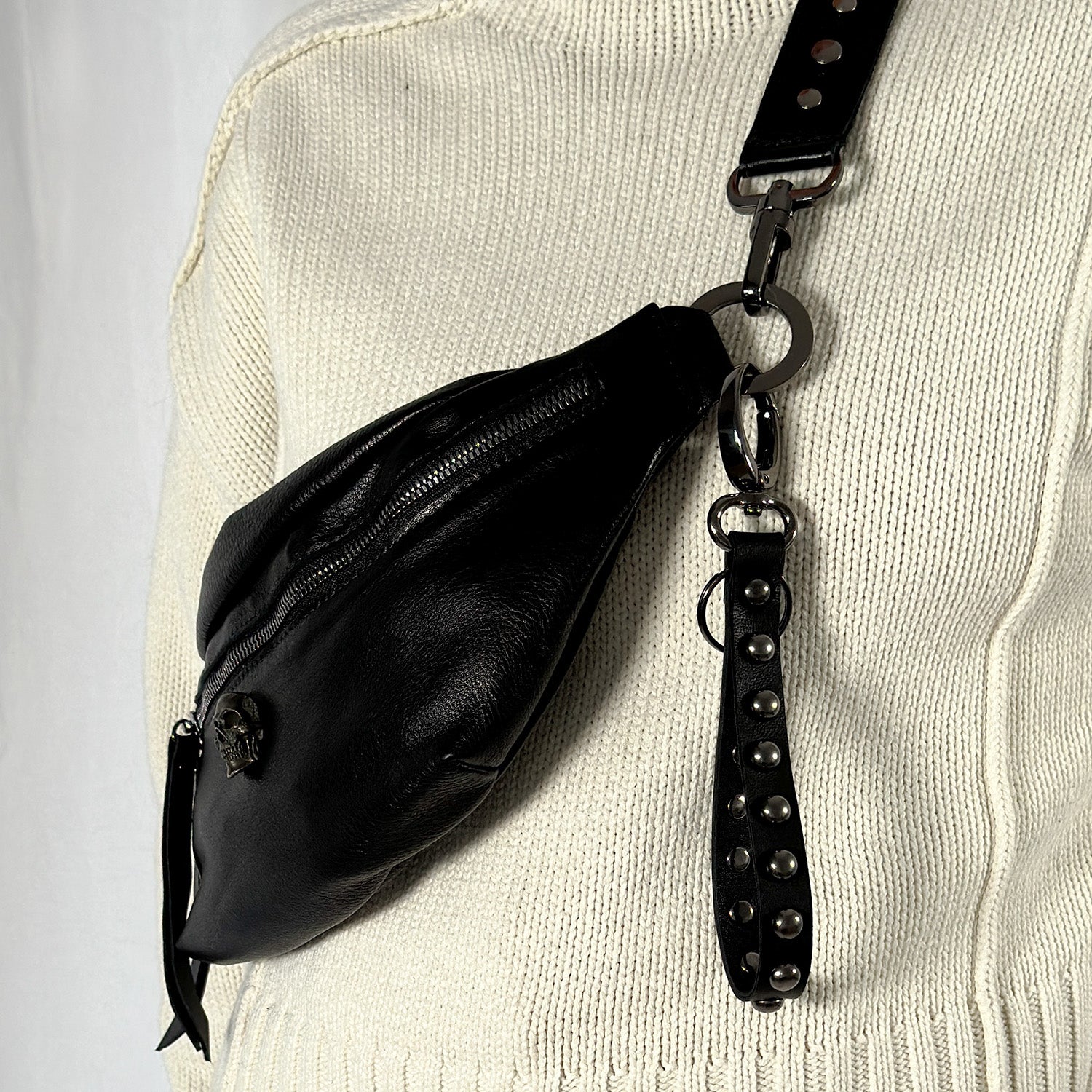 The KEY RING - BLACK STUDDED from evokewomen, a black leather crossbody bag featuring silver hardware and a studded key ring strap, worn over a beige knit sweater.
