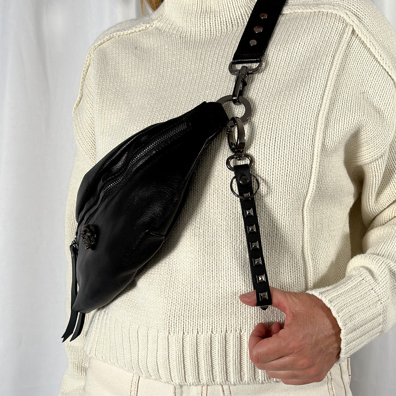 A person is wearing a cream sweater and holding an evokewomen crossbody bag called "KEY RING - BLACK PYRAMID RIVETS," featuring a studded strap with unique design elements, against a white background.