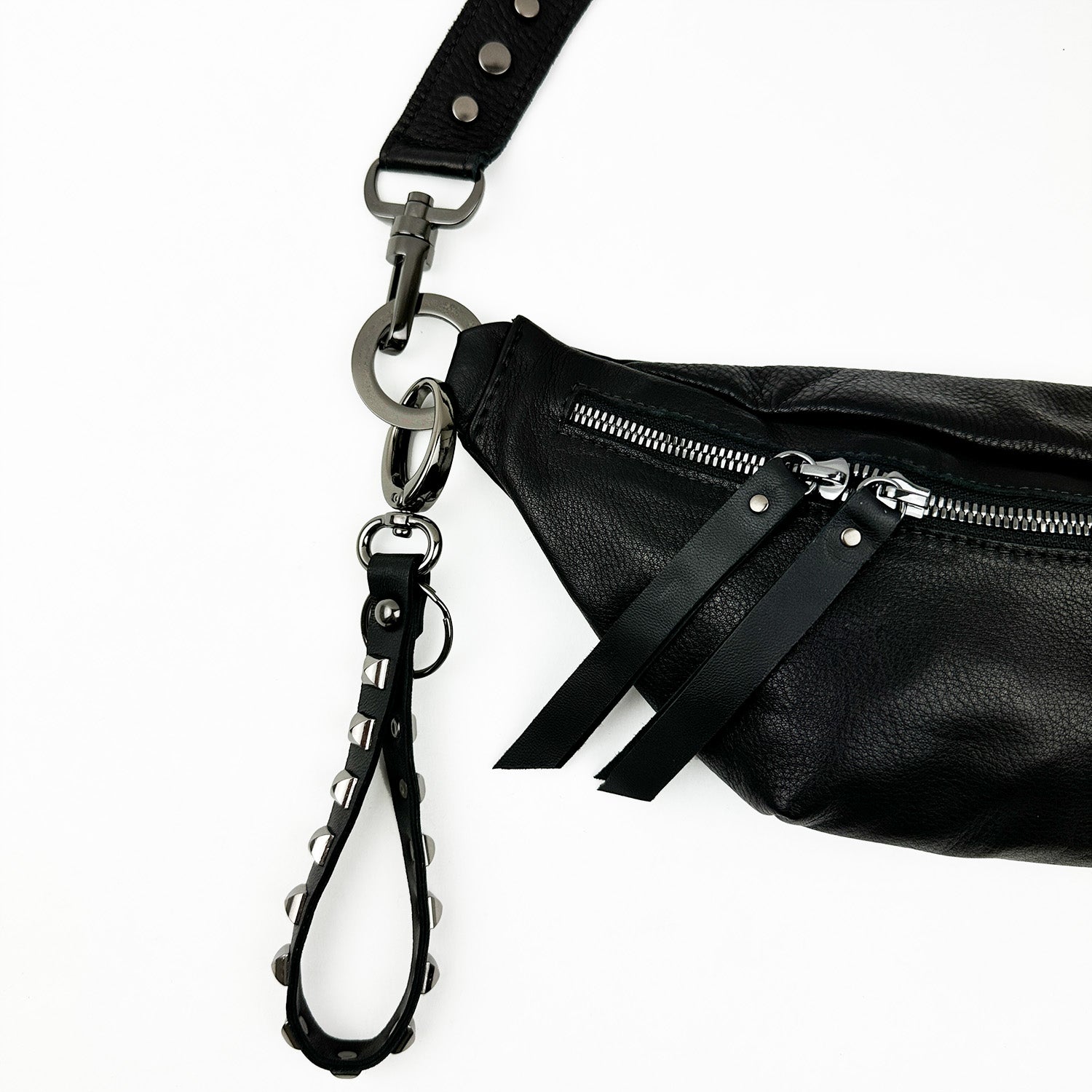 The KEY RING - BLACK PYRAMID RIVETS by evokewomen is a chic black leather clutch bag enhanced with metal zippers and decorative pull straps. It features a detachable studded wrist strap connected by a metal clasp, along with a unique accessory for carrying keys using the stylish studded key ring strap.