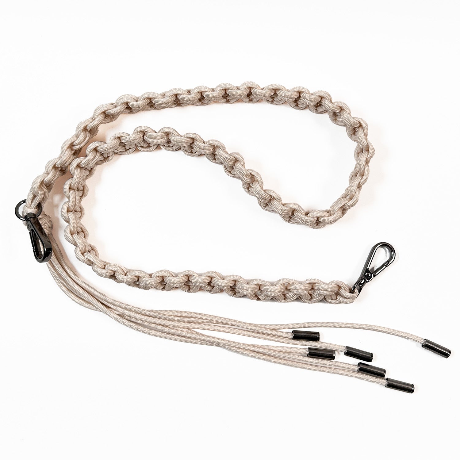 This image shows an intricately woven paracord bag strap in a light beige color. The cords are knotted in a flat, decorative braid style and finished with gunmetal swivel snap hooks for attaching to a bag. One strap end features additional paracord strands forming a tassel.