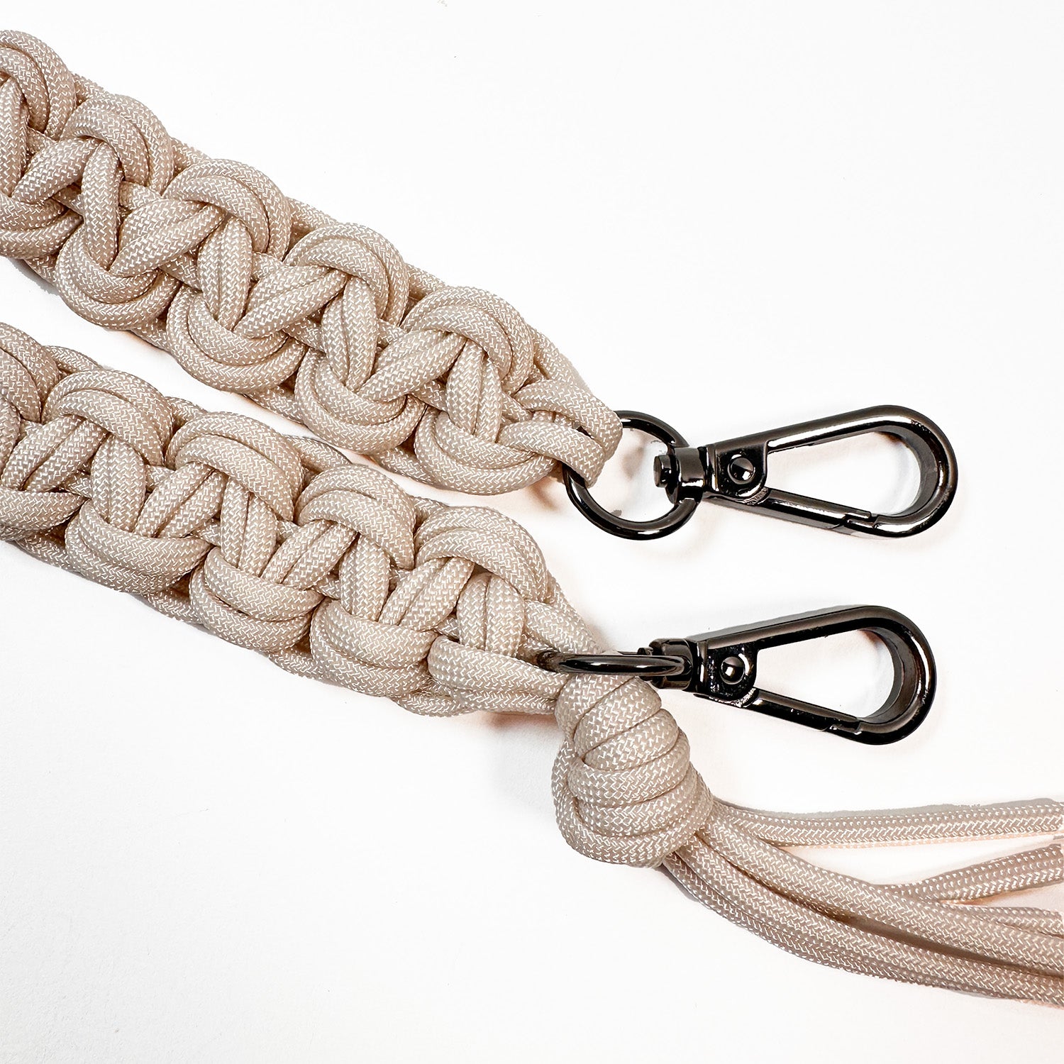 This image shows a close-up of two intricately woven paracord bag straps in a light beige color. The cords are knotted in a flat, decorative braid style and finished with gunmetal swivel snap hooks for attaching to a bag. One strap end features additional paracord strands forming a tassel.