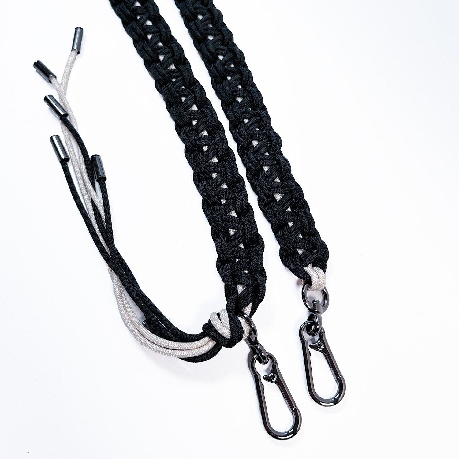 This image shows a close-up of two intricately woven paracord bag straps in midnight blue and off white. The cords are knotted in a flat, decorative braid style and finished with gunmetal swivel snap hooks for attaching to a bag. One strap end features additional paracord strands forming a tassel.