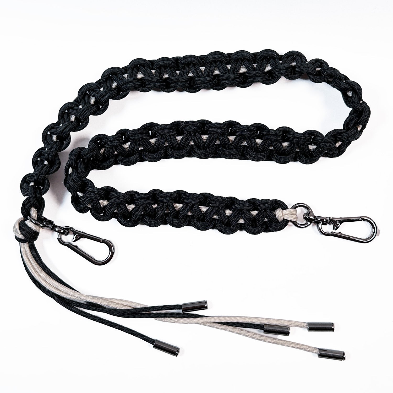 This image shows an intricately woven paracord bag strap in midnight blue and off white. The cords are knotted in a flat, decorative braid style and finished with gunmetal swivel snap hooks for attaching to a bag. One strap end features additional paracord strands forming a tassel.
