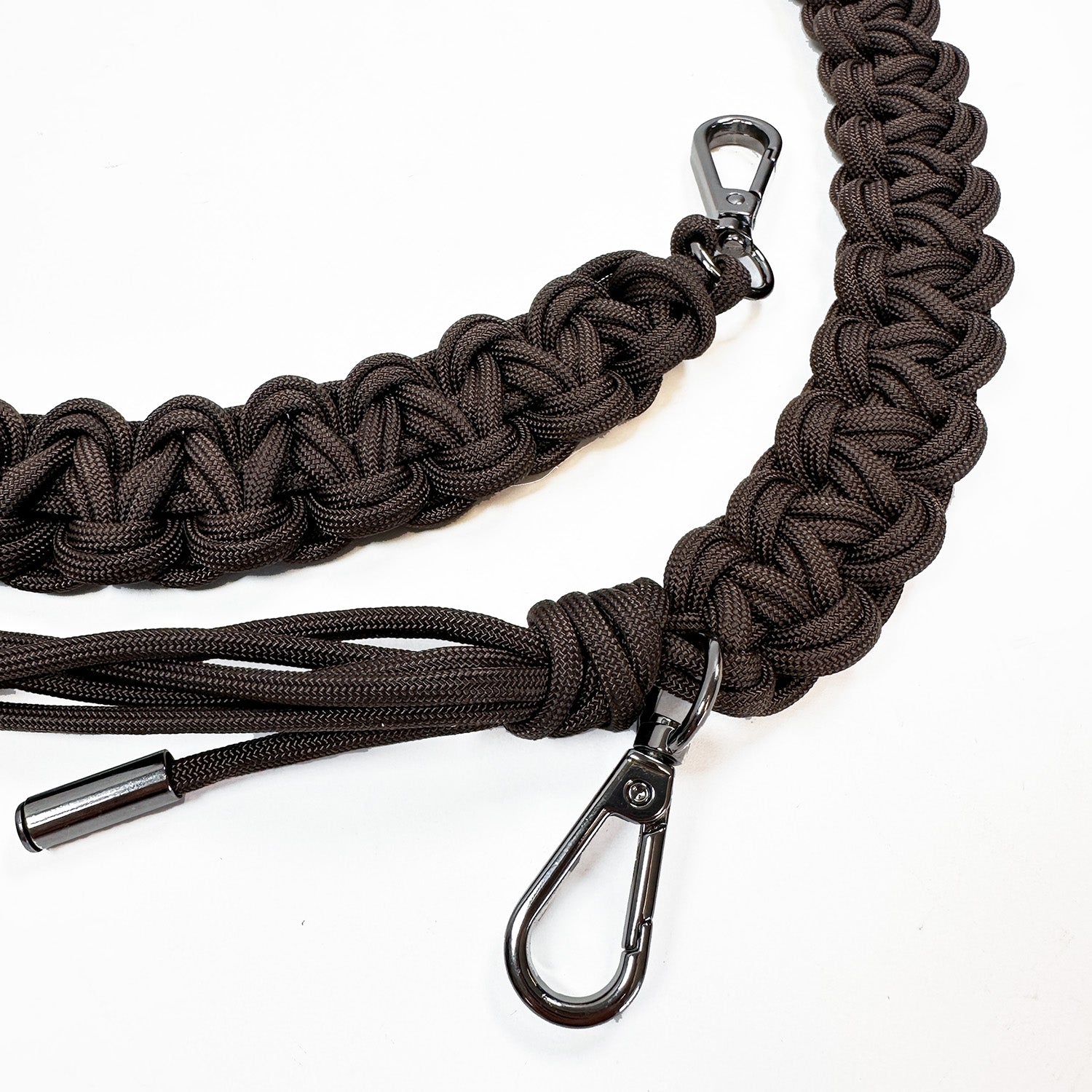 This image shows a close-up of two intricately woven paracord bag straps in dark brown. The cords are knotted in a flat, decorative braid style and finished with gunmetal swivel snap hooks for attaching to a bag. One strap end features additional paracord strands forming a tassel.