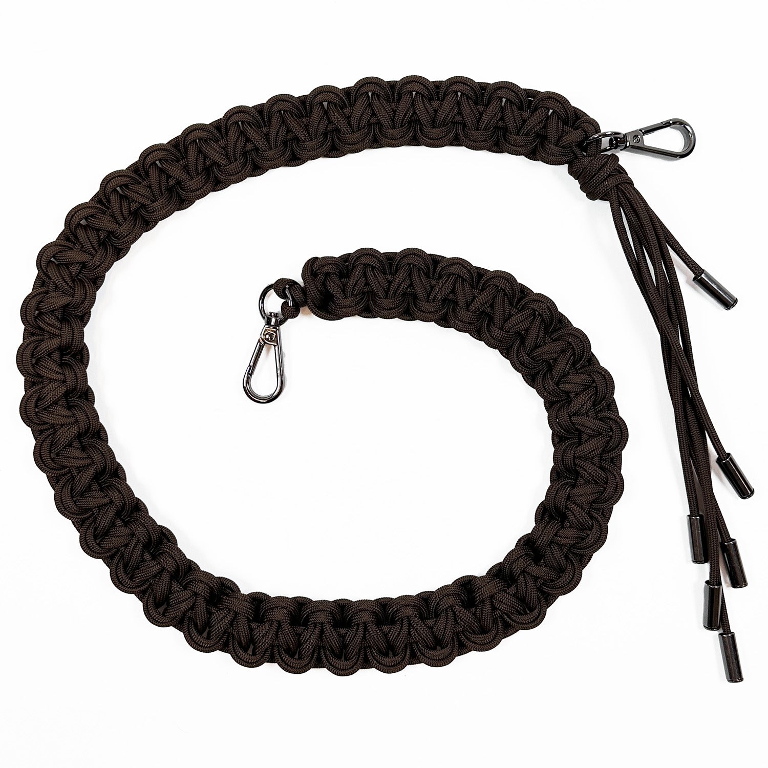 This image shows an intricately woven paracord bag strap in dark brown. The cords are knotted in a flat, decorative braid style and finished with gunmetal swivel snap hooks for attaching to a bag. One strap end features additional paracord strands forming a tassel.