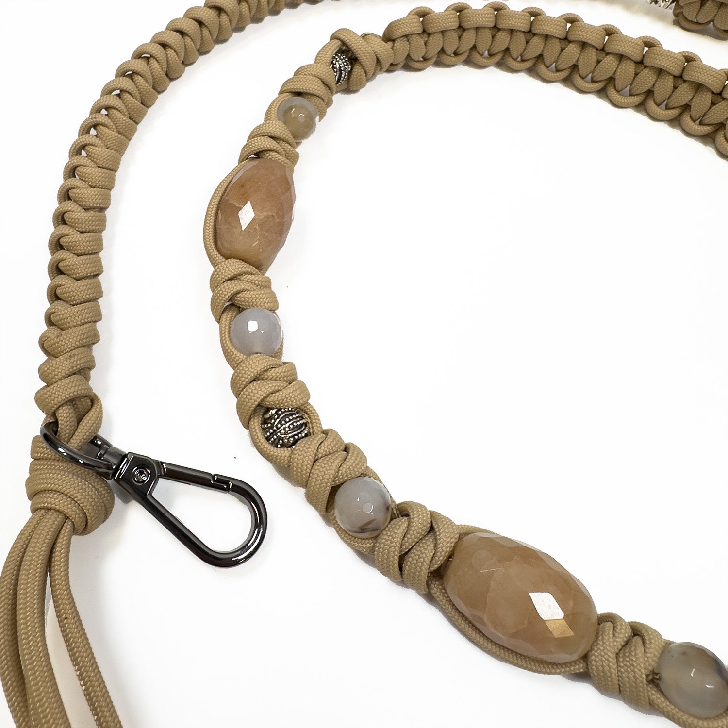 A detailed view of the KNOTTED SHOULDER STRAP - CAMEL by evokewomen showcases its beige braided design embellished with decorative stones and antique silver metal beads. It includes a black metal clip for secure fastening.