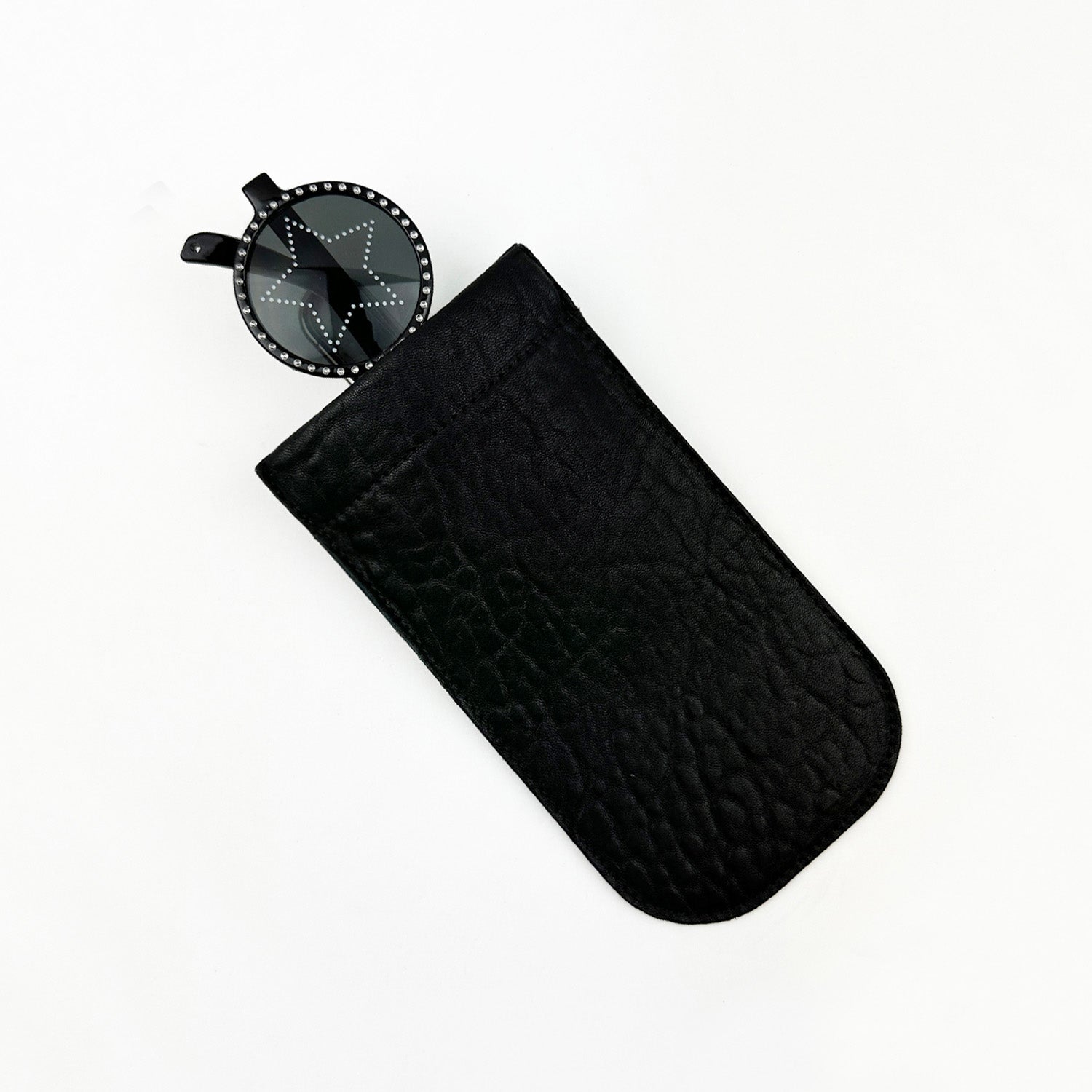 The SUN-GLASS POUCH - BLACK BUBBLY by evokewomen is a black leather pouch with a squeeze frame closure, showcasing a partially visible round tag with a star pattern at the top, perfect for protecting eyewear against a plain white background.