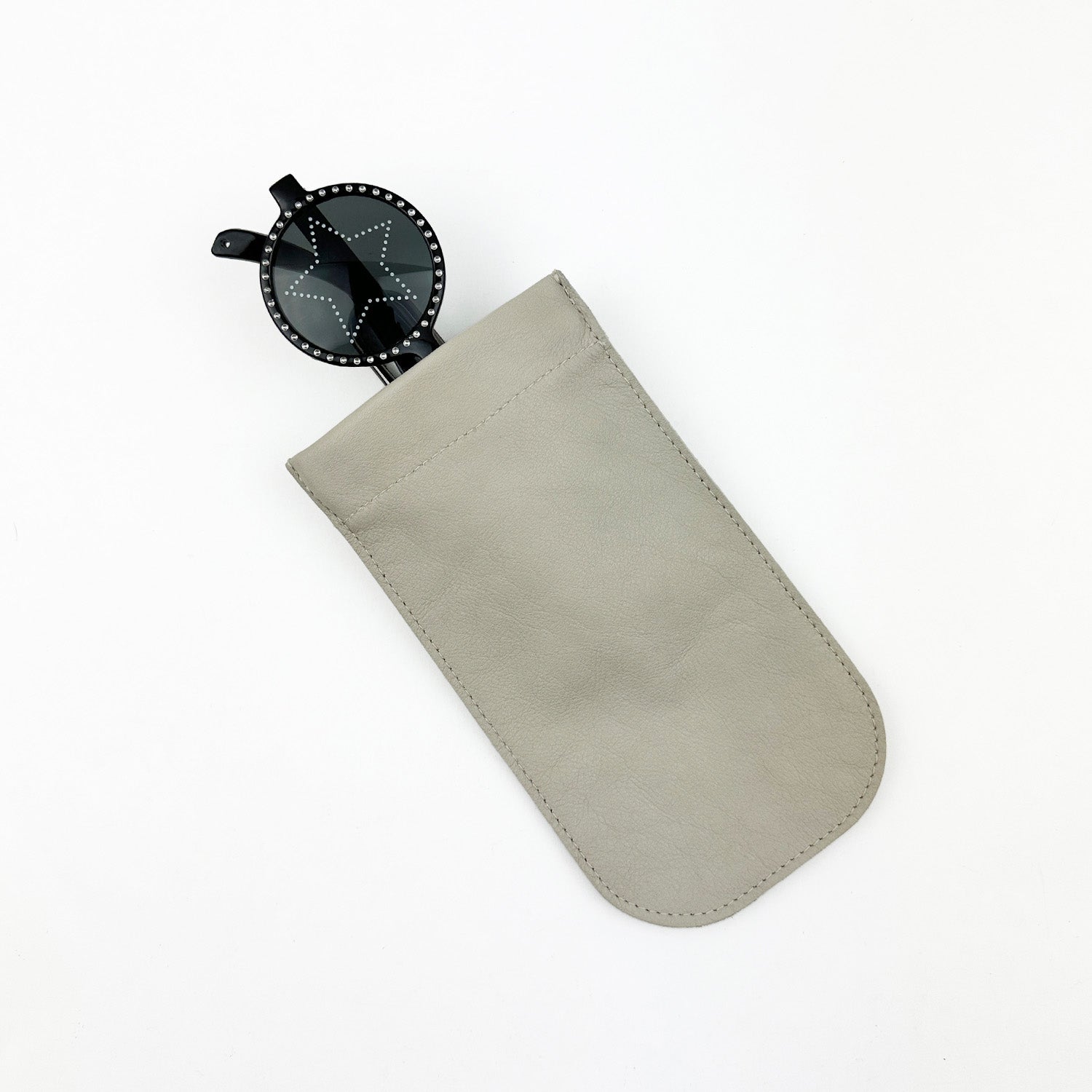 A pair of black star-shaped sunglasses rests in a sleek leather evokewomen SUN-GLASS POUCH - PUTTY, featuring a high-quality gunmetal spring hook, set against a white background.