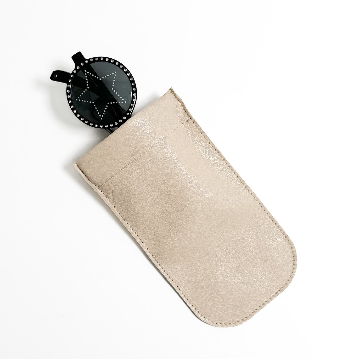 A round, black object featuring a star design is partially placed inside an evokewomen SUN-GLASS POUCH in almond leather with a squeeze frame closure, all set against a white background.
