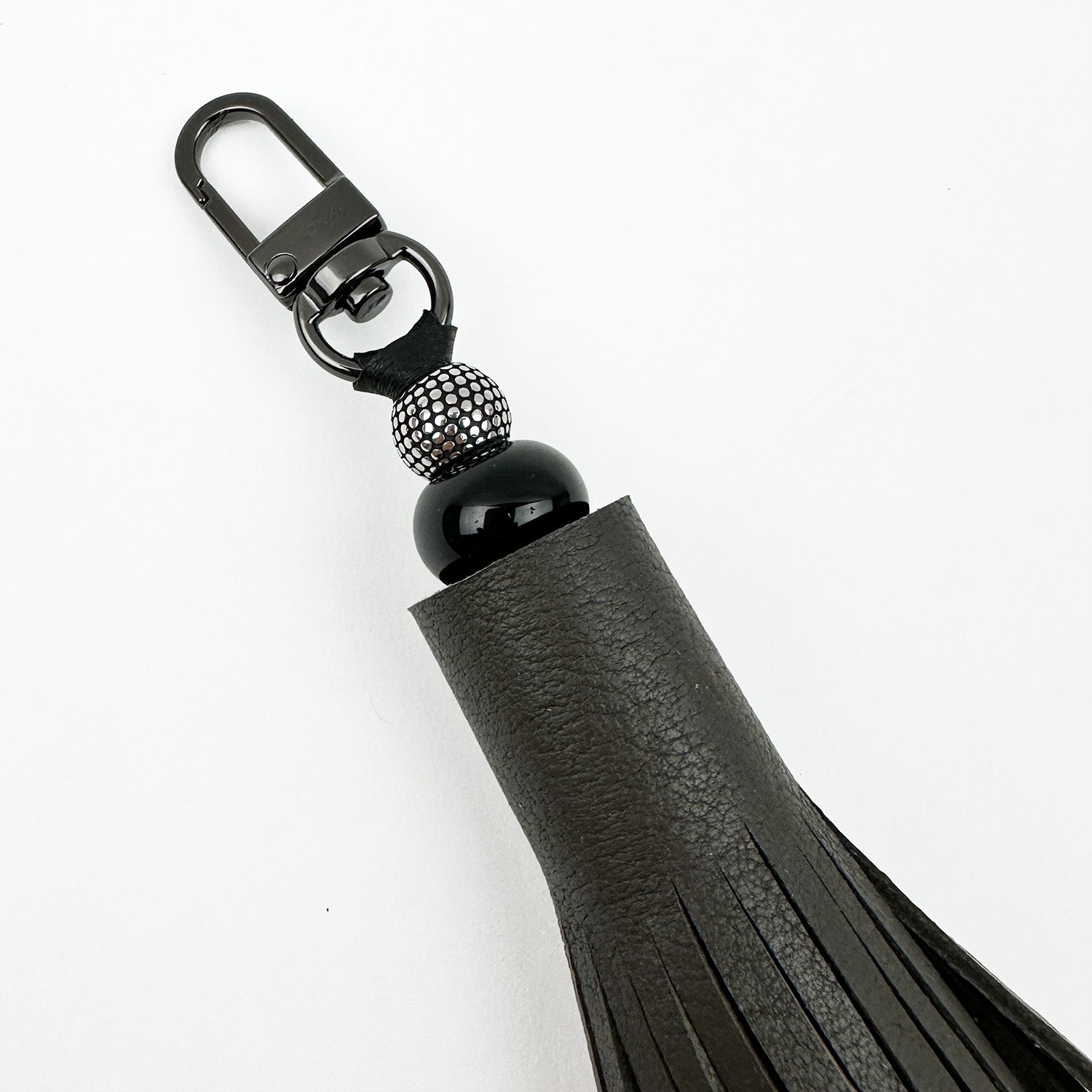 Close-up of the evokewomen TASSEL - OLIVE-GREY keychain in black leather with tiered tassels, a gunmetal swivel hook, and beaded accents set against a white background.