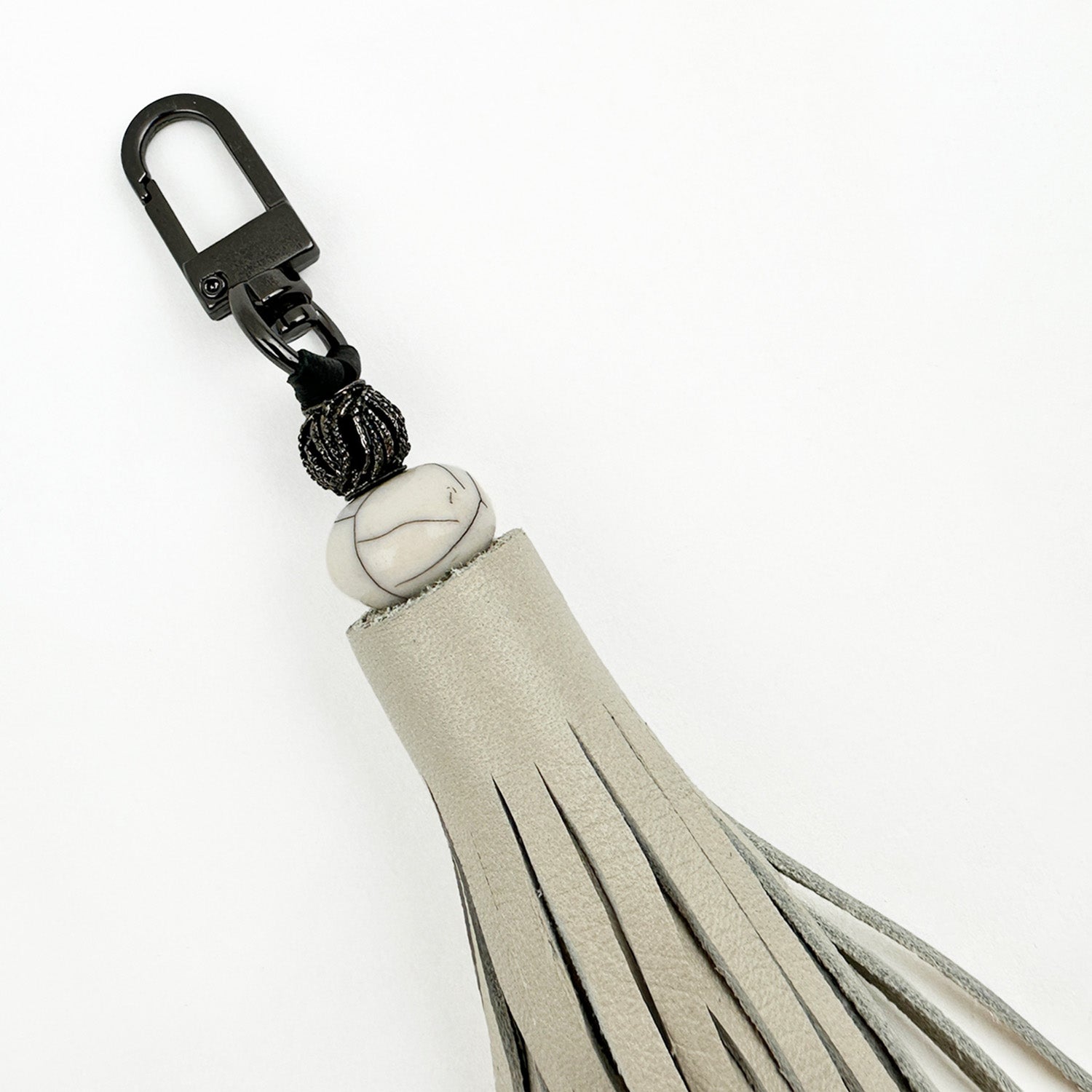 Introducing the TASSEL - PUTTY by evokewomen: a uniquely designed gray tiered tassel keychain, featuring a gunmetal accessory clip, a distinctive marbled bead, and a black textured bead above the tassel.