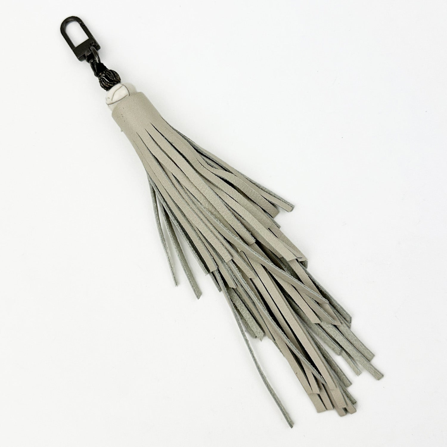 The TASSEL - PUTTY from evokewomen is a gray leather tiered tassel keychain featuring a gunmetal accessory and metal clip attachment, showcased against a plain white background.