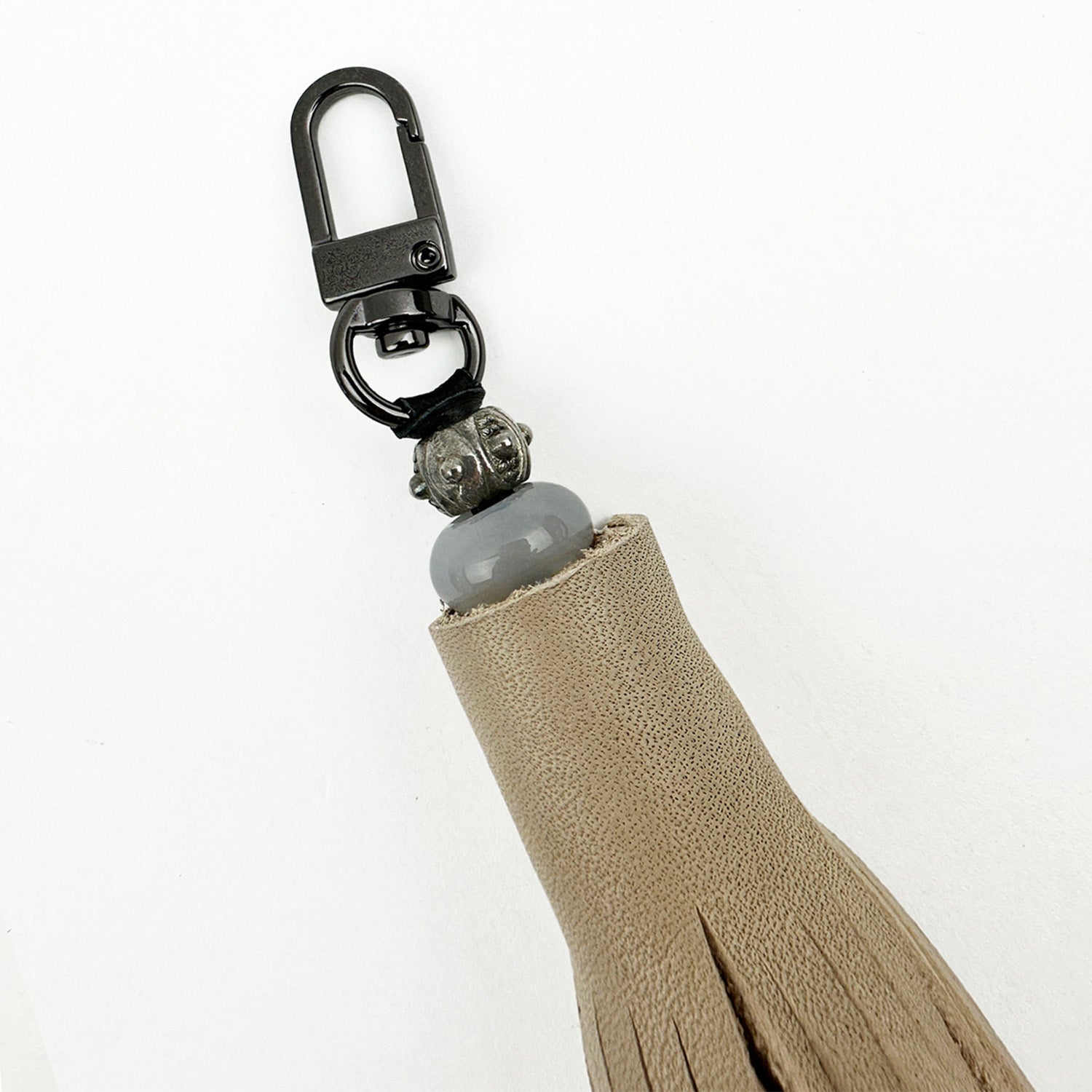 Introducing the TASSEL - SAND by evokewomen: a one-of-a-kind tiered tassel keychain in beige, highlighted with a dark gunmetal clasp and a round gray bead, all perfectly showcased against a white background.