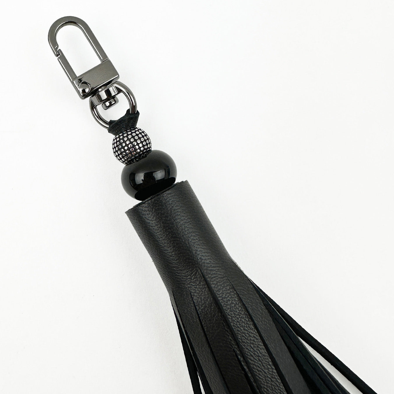 The TASSEL - BLACK, from evokewomen, is a black leather keychain showcasing a distinctive tiered tassel design, adorned with a decorative bead and equipped with a durable gunmetal swivel hook.