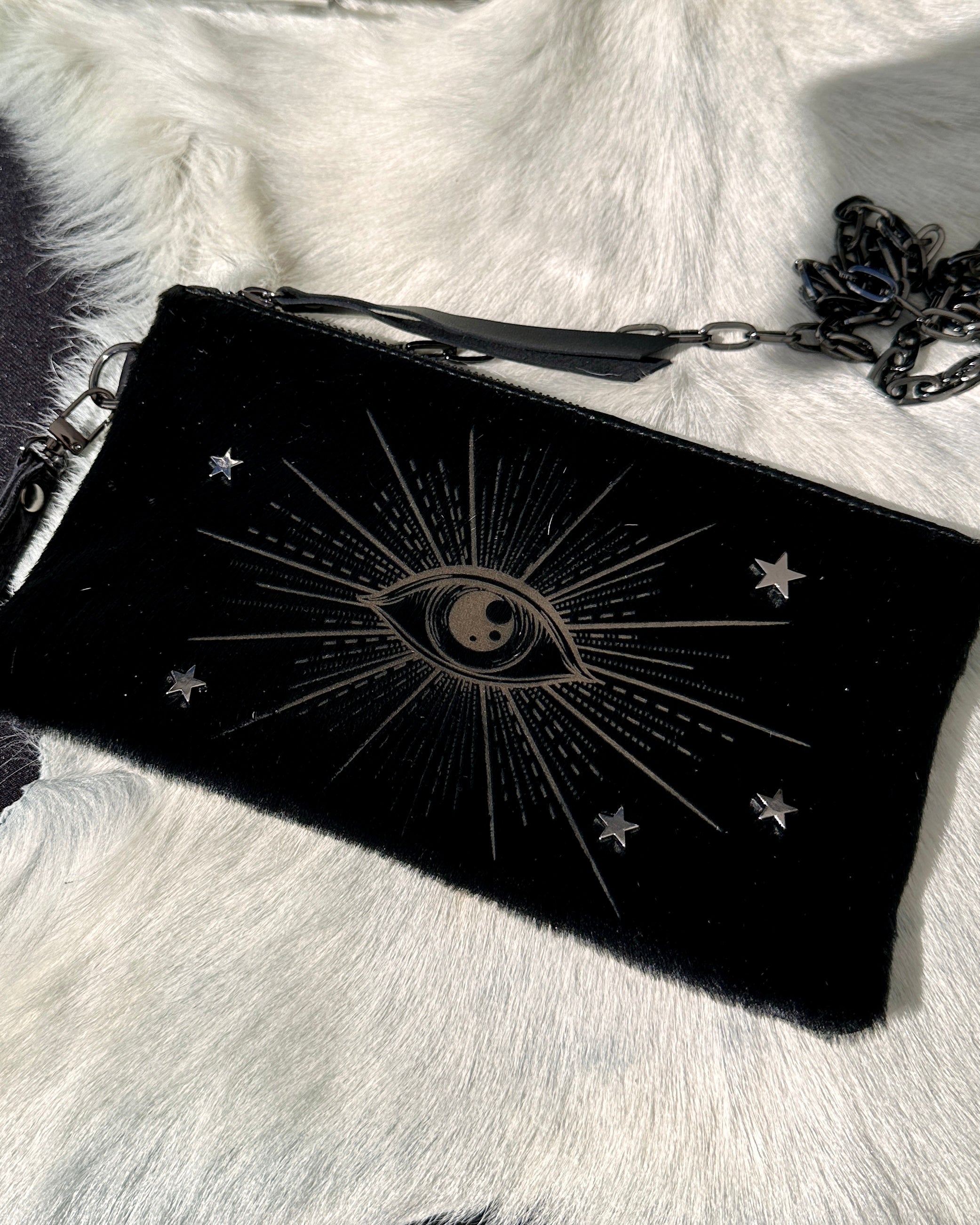 The VICTORIA - black - laser eye wristlet from evokewomen, adorned with a chain and an eye design encircled by stars and radiating lines, is elegantly displayed on a white fur surface.