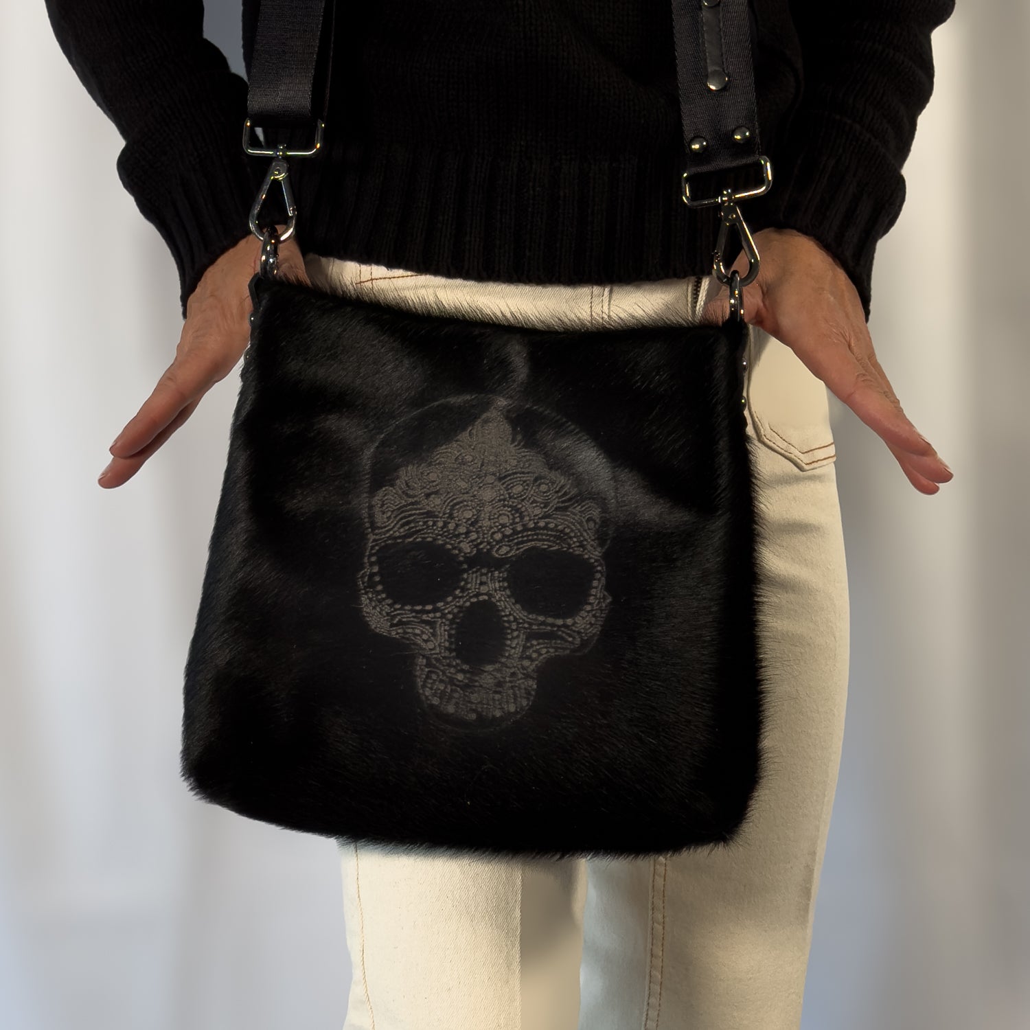 A person is carrying the ENYA - BLACK LASER SKULL crossbody bag by evokewomen, paired with a black sweater and light-colored pants.