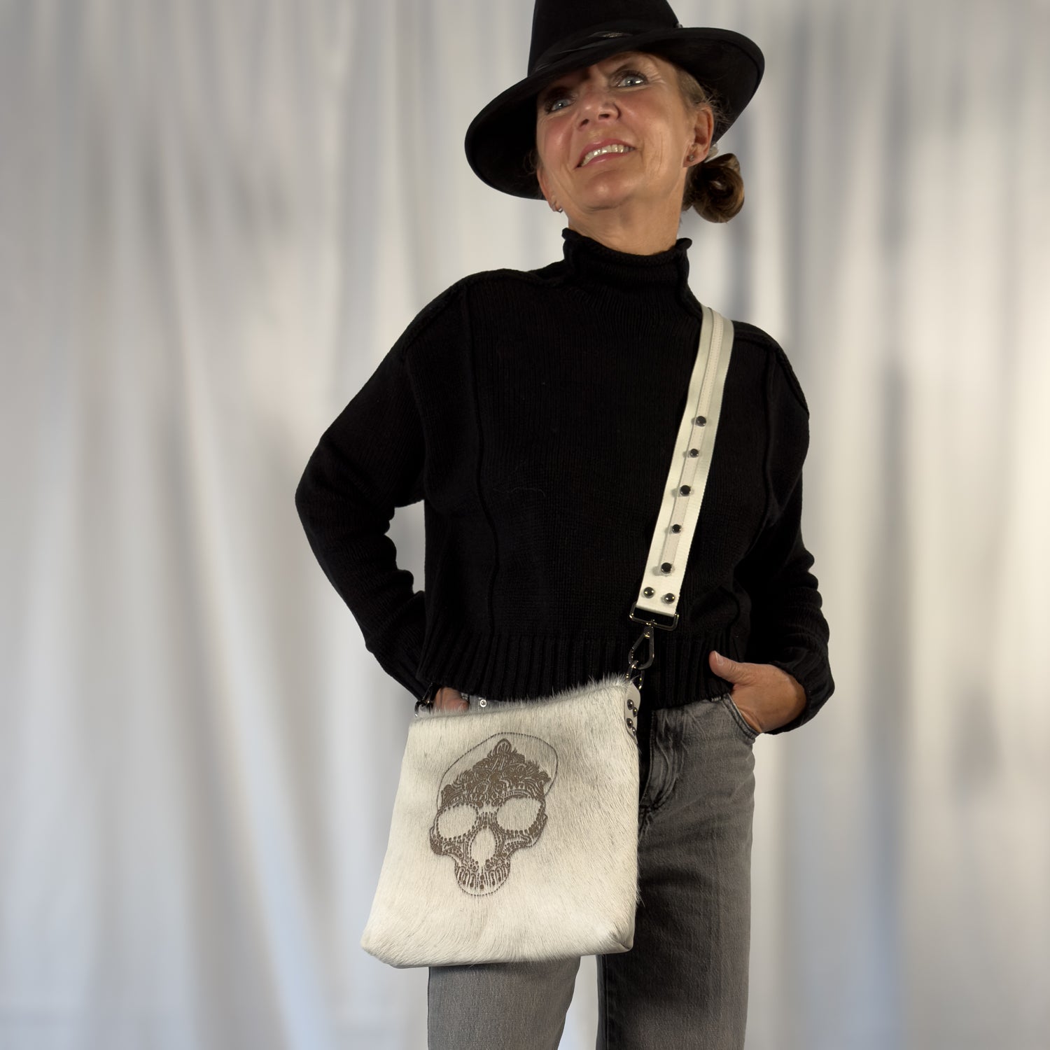An individual wearing a black hat and sweater smiles with hands in pockets, carrying the ENYA - OFF-WHITE LASER SKULL bag by evokewomen, featuring an eye-catching skull laser design. The background is plain.