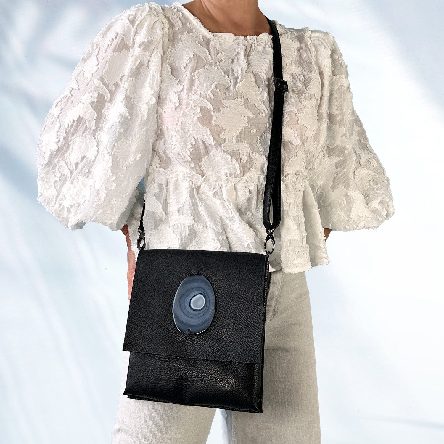 Woman wearing a textured white lace blouse and light gray jeans, carrying a black leather crossbody bag with an oval blue-gray agate stone on the front flap, standing against a light blue background.