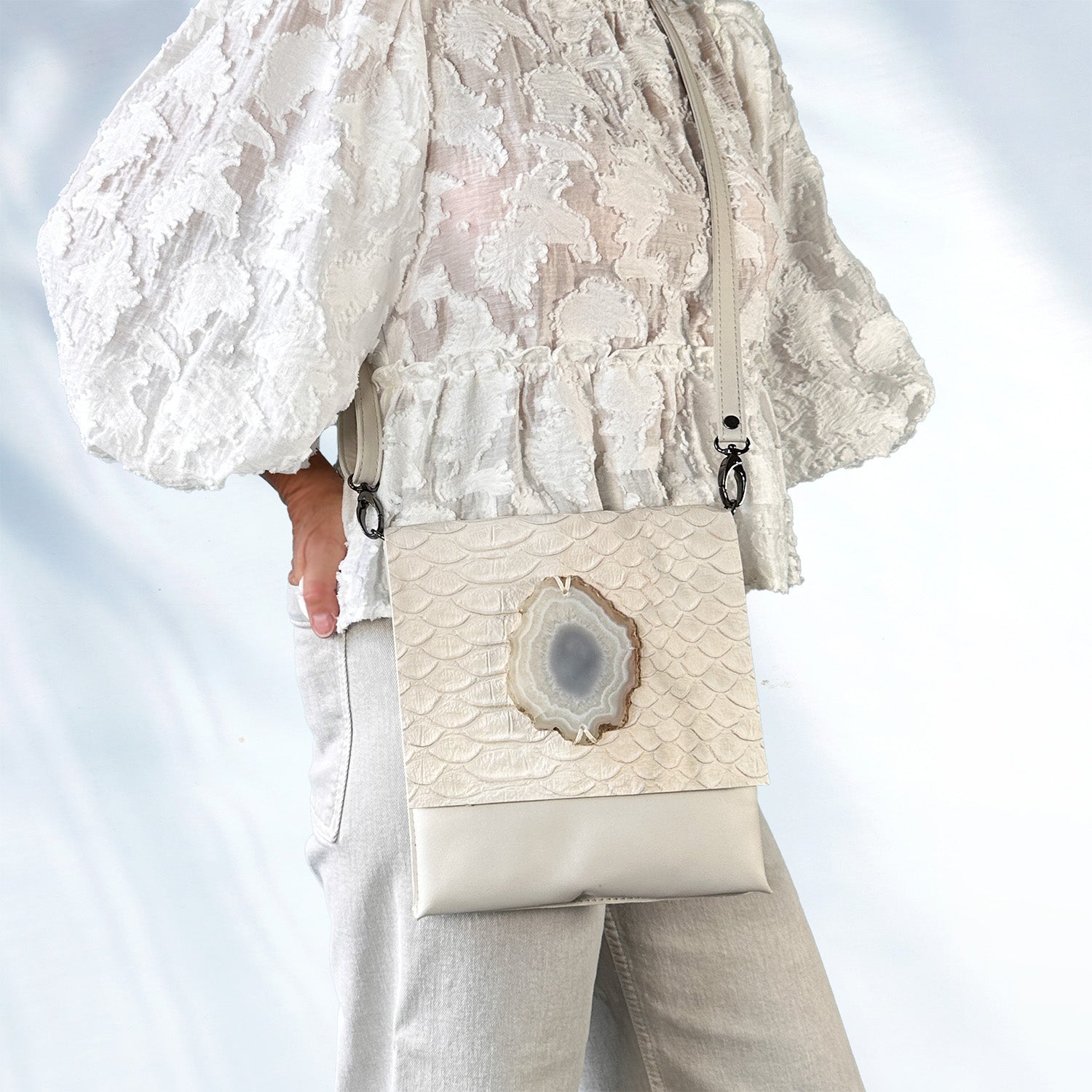 Woman wearing a textured white lace blouse and light gray jeans, carrying a cream-colored crossbody bag made of embossed leather with a large oval agate stone on the front flap, standing against a soft light blue background.