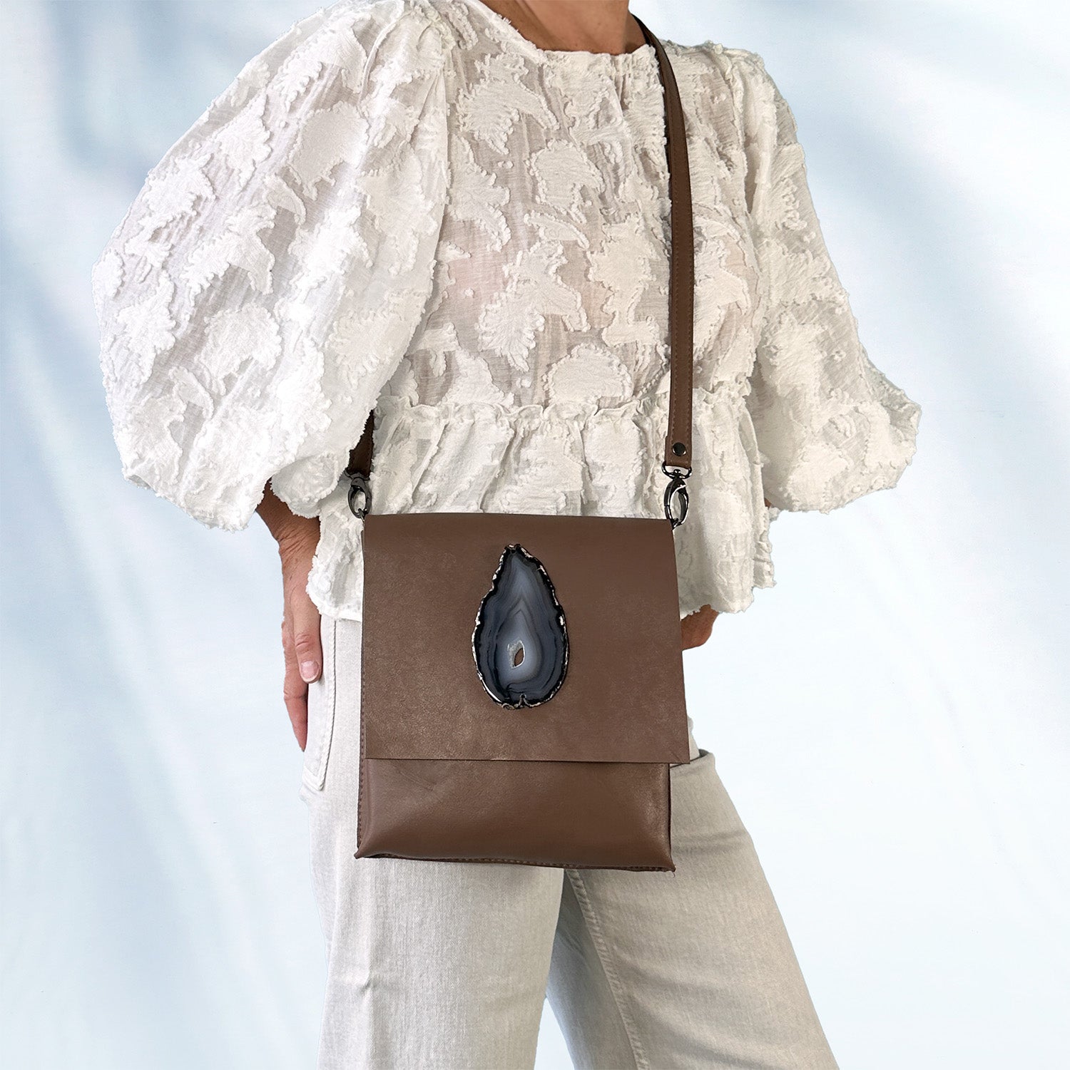 Woman wearing a textured white lace blouse and light gray jeans, carrying a black leather crossbody bag with an teardrop shaped black-gray agate stone on the front flap, standing against a light blue background.