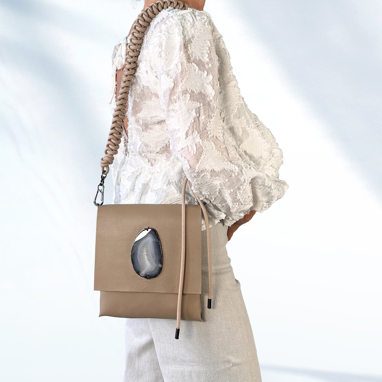Woman in a white textured lace blouse and light gray pants wearing a taupe leather crossbody bag with a teardrop-shaped agate stone on the flap and an edgy, knotted shoulder strap, standing against a soft light blue background.