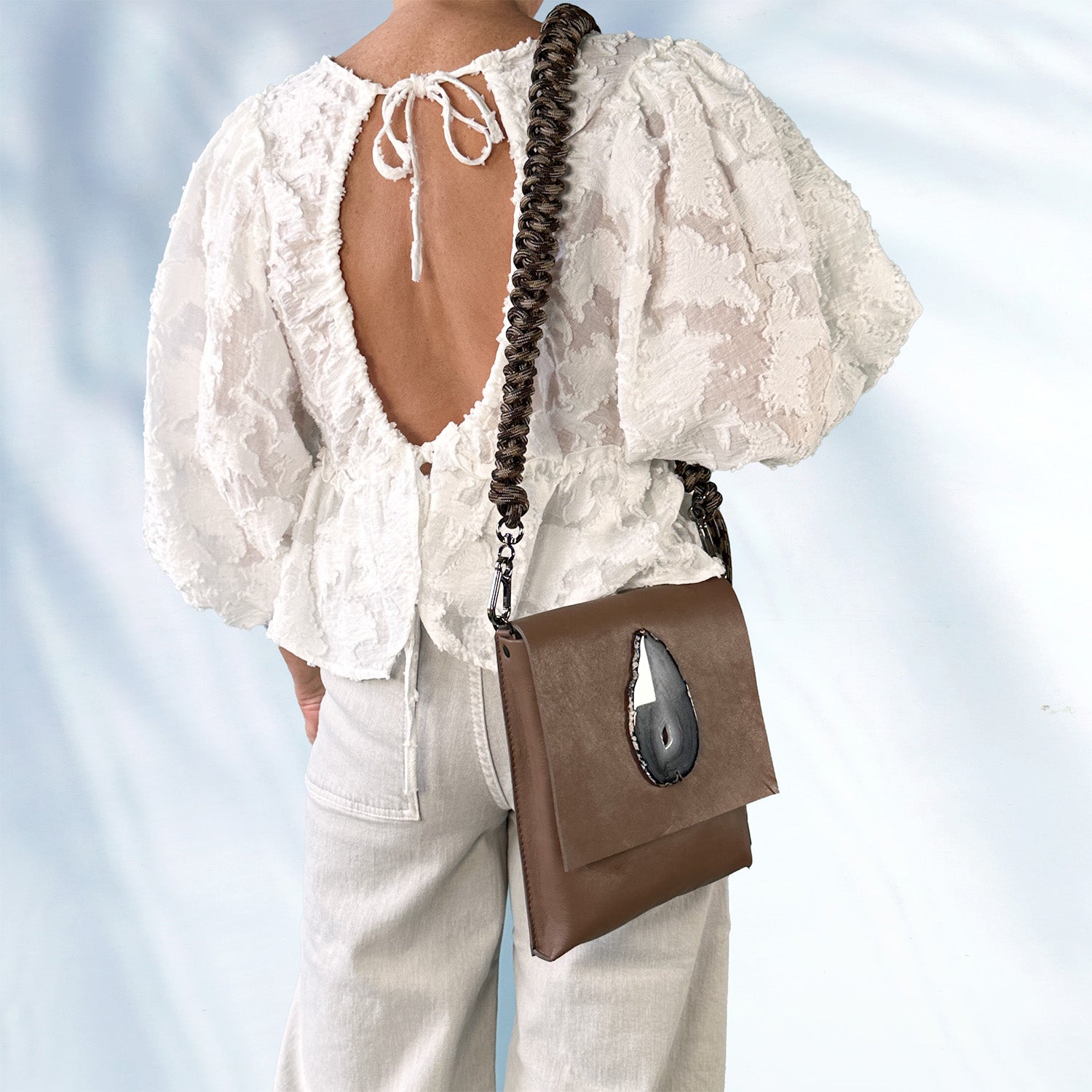 Woman in a white textured lace blouse and light gray pants wearing a mocha brown leather crossbody bag with a teardrop-shaped agate stone on the flap and an edgy, knotted shoulder strap, standing against a soft light blue background.