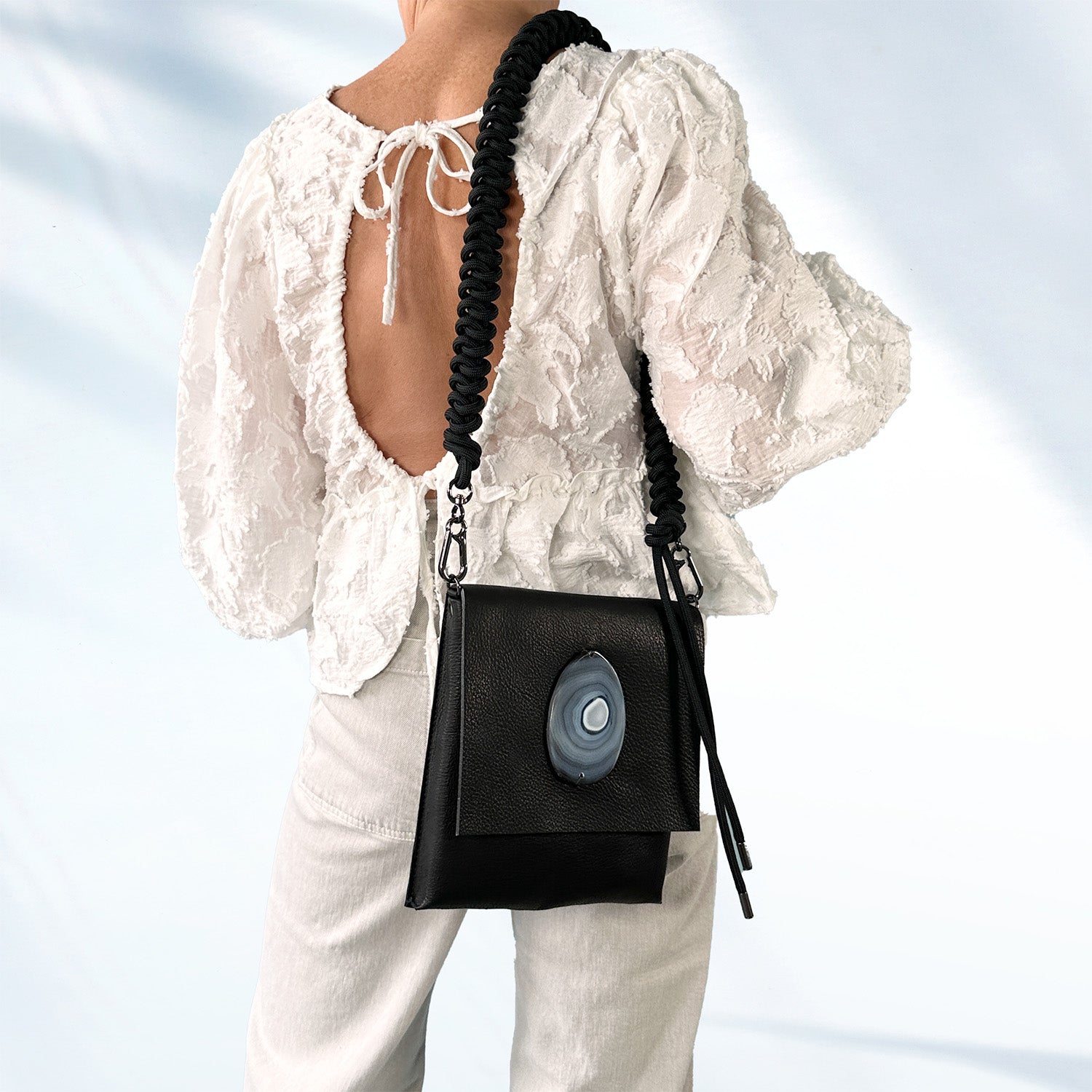 Woman in a white textured lace blouse and light gray pants wearing a black leather crossbody bag with an oval -shaped agate stone on the flap and an edgy, knotted shoulder strap, standing against a soft light blue background.