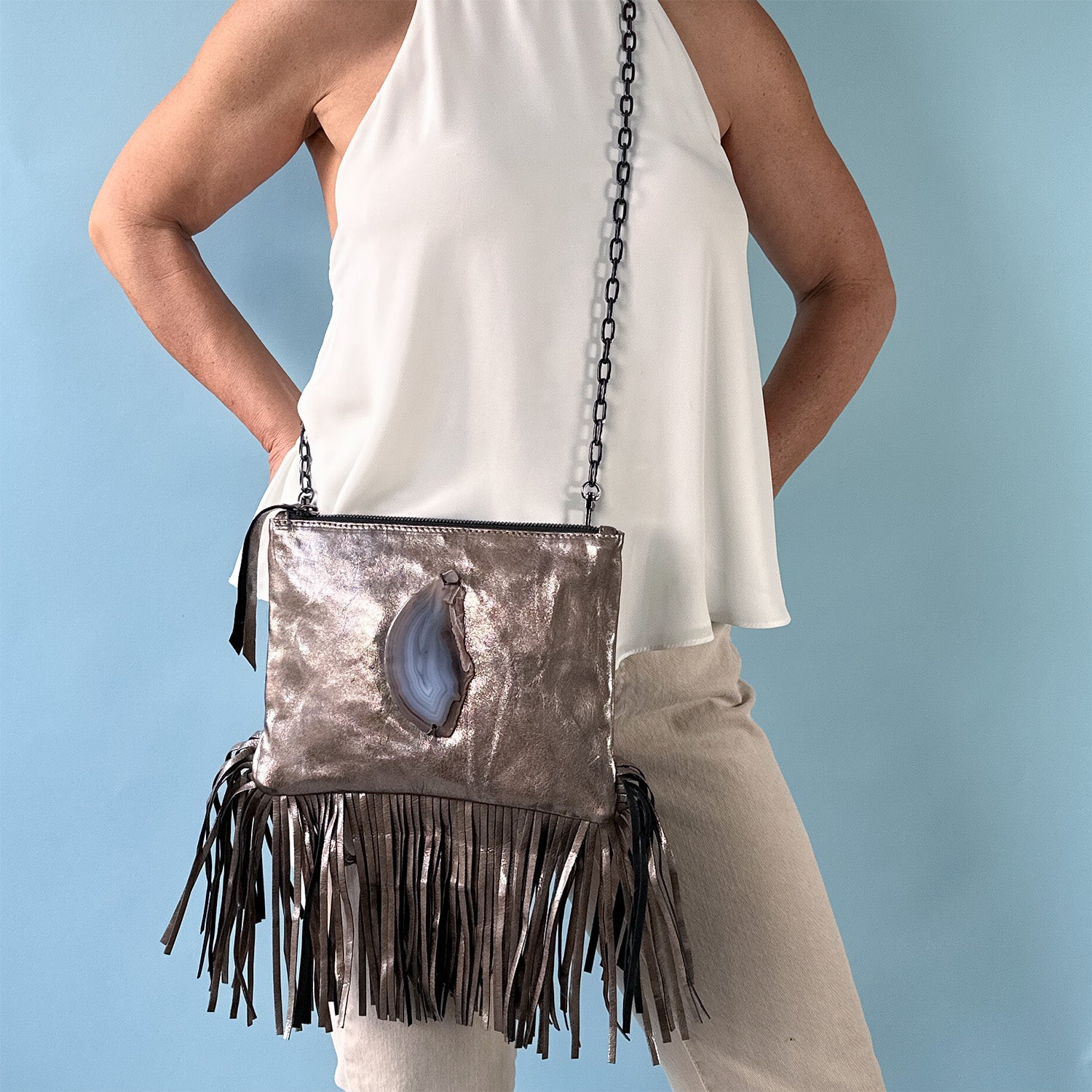 Woman wearing a sleeveless white halter top and beige pants, modeling a brown metallic leather crossbody bag with long fringe detailing and a teardrop-shaped agate stone accent on the front, against a light blue background.