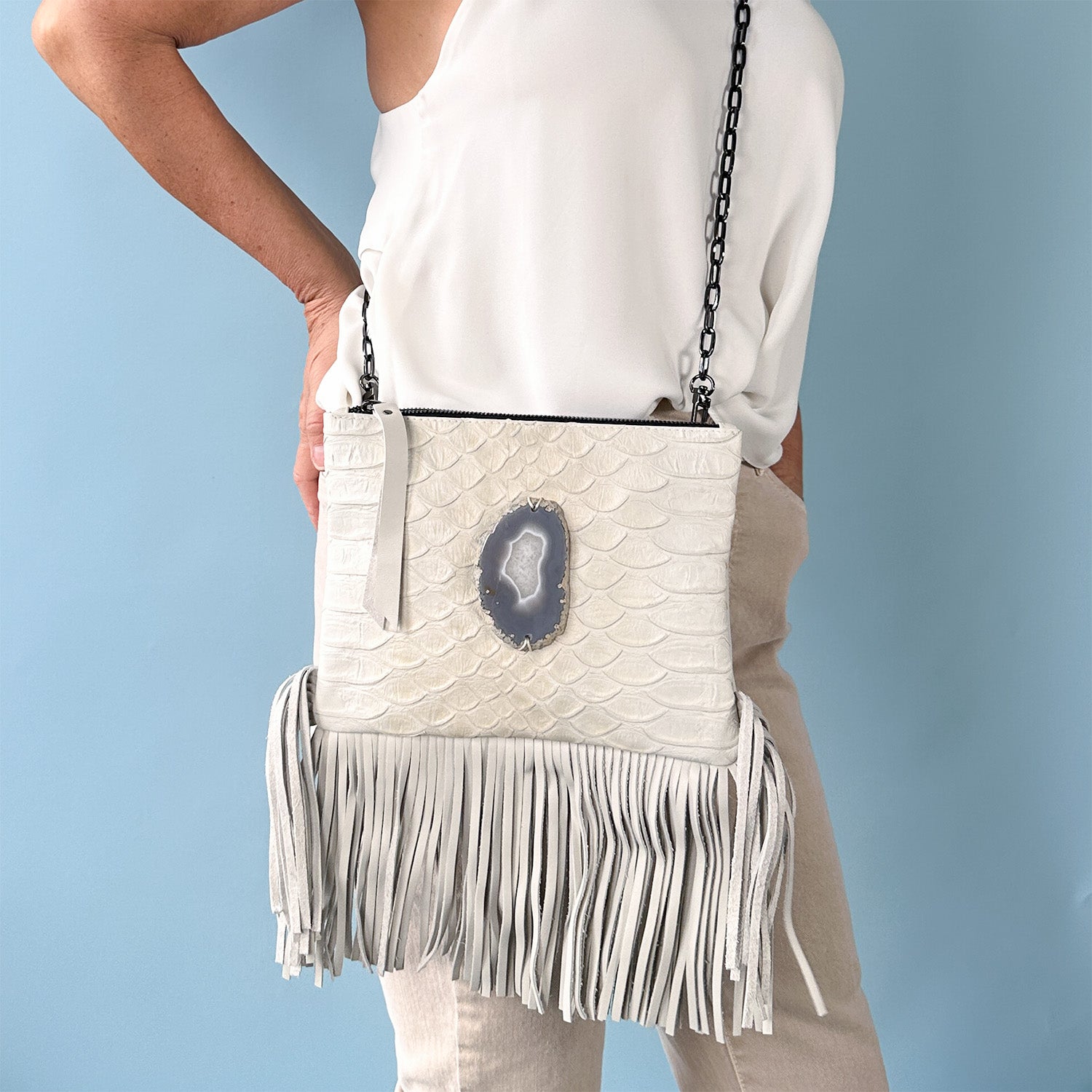 Woman wearing a sleeveless white halter top and beige pants, modeling a off-white leather crossbody bag with a scale-textured surface, long fringe at the bottom, and a large oval-shaped gray agate stone accent in the center, against a light blue background.