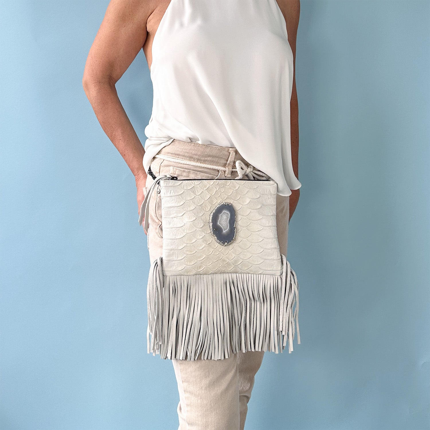 Woman wearing a sleeveless white halter top and beige pants, modeling a off-white leather crossbody bag with a scale-textured surface, long fringe at the bottom, and a large oval-shaped gray agate stone accent in the center, worn around the waist as a belt bag against a light blue background.