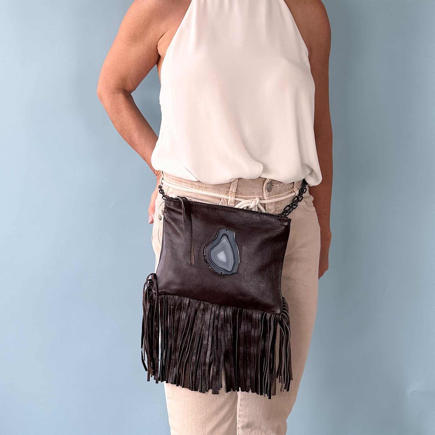 Woman wearing a sleeveless white halter top and beige pants, modeling a brown leather fringe bag with a teardrop-shaped agate stone accent, worn around the waist as a belt bag, against a light blue background.