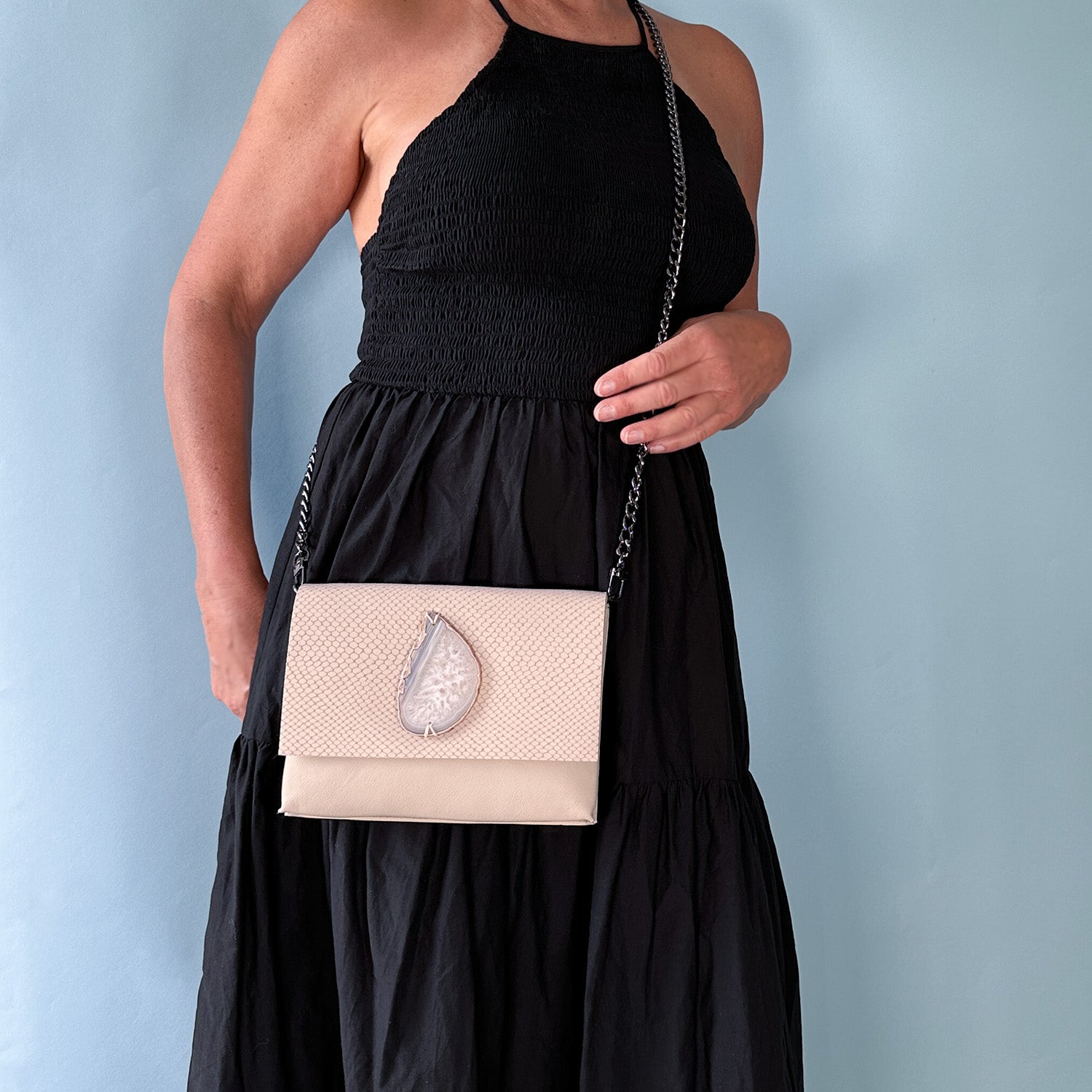 Woman wearing a sleeveless black dress with a blush-toned leather crossbody bag. The bag features a textured flap, a teardrop-shaped light agate stone centerpiece, and a gunmetal chain strap. Set against a soft blue background.