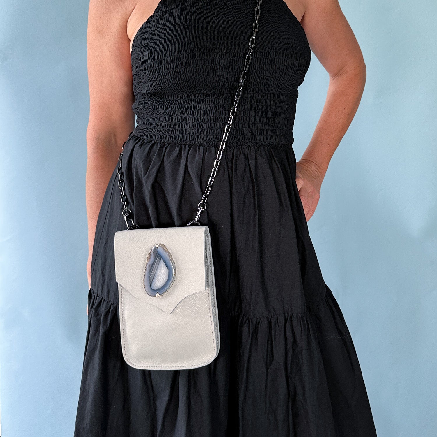 Woman wearing a sleeveless black dress with a light grey leather crossbody bag. The bag features a teardrop-shaped blue agate stone and a gunmetal chain strap. Set against a soft blue background.