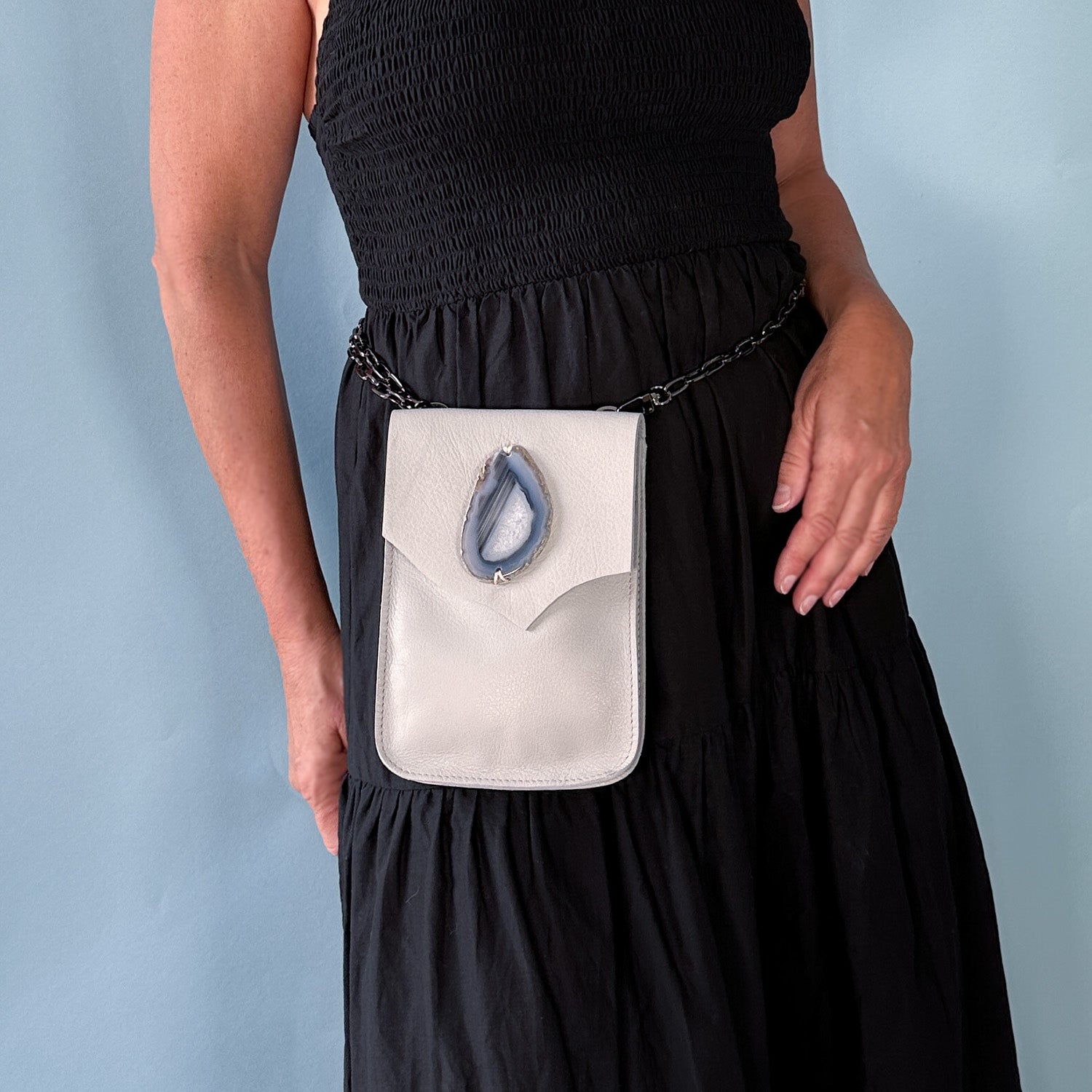 Woman wearing a sleeveless black dress with a light grey leather bag around the waist. The bag features a teardrop-shaped blue agate stone and a gunmetal chain strap. Set against a soft blue background.