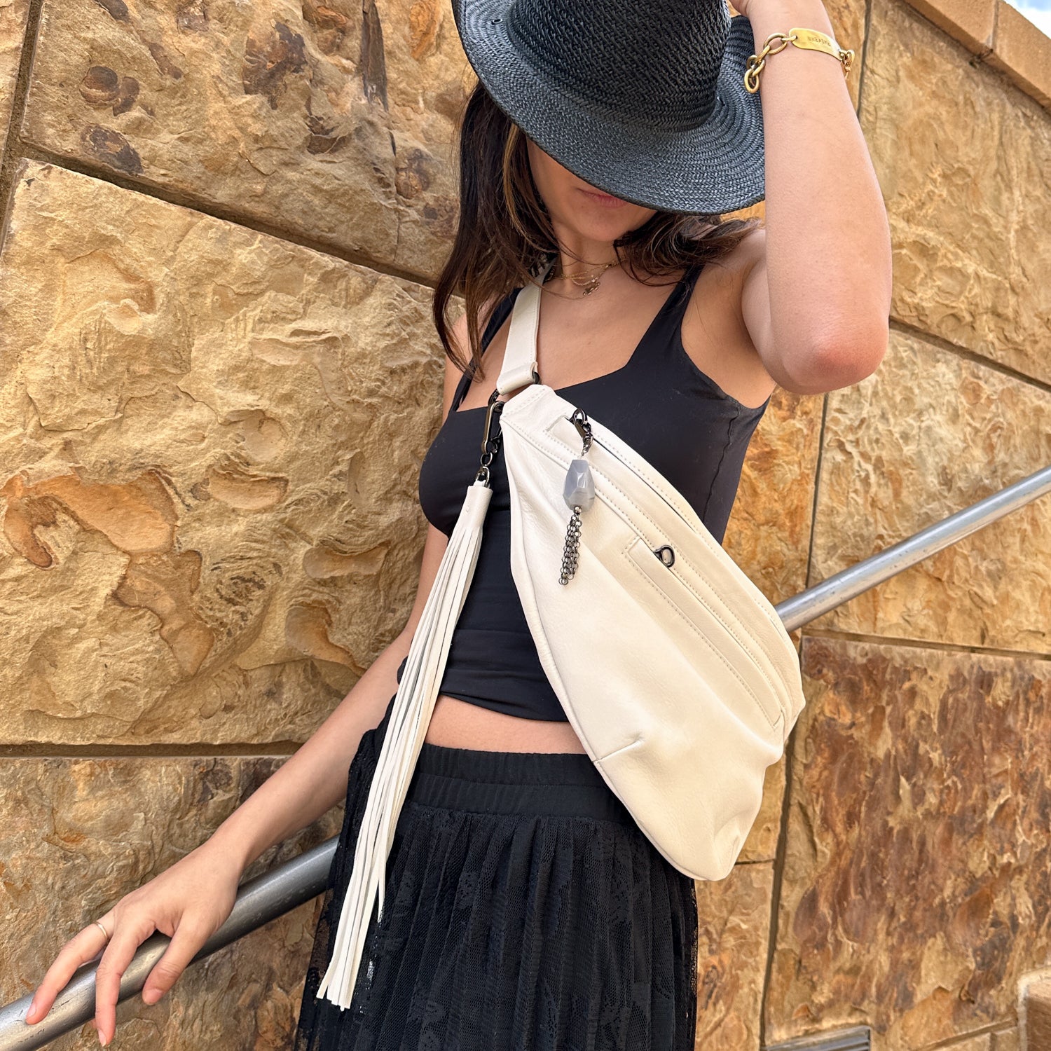 A person in a stylish black top and hat holds the brim, wearing the large and versatile Sienna - pearl white & tassel bag by evokewomen, standing against a textured stone wall.
