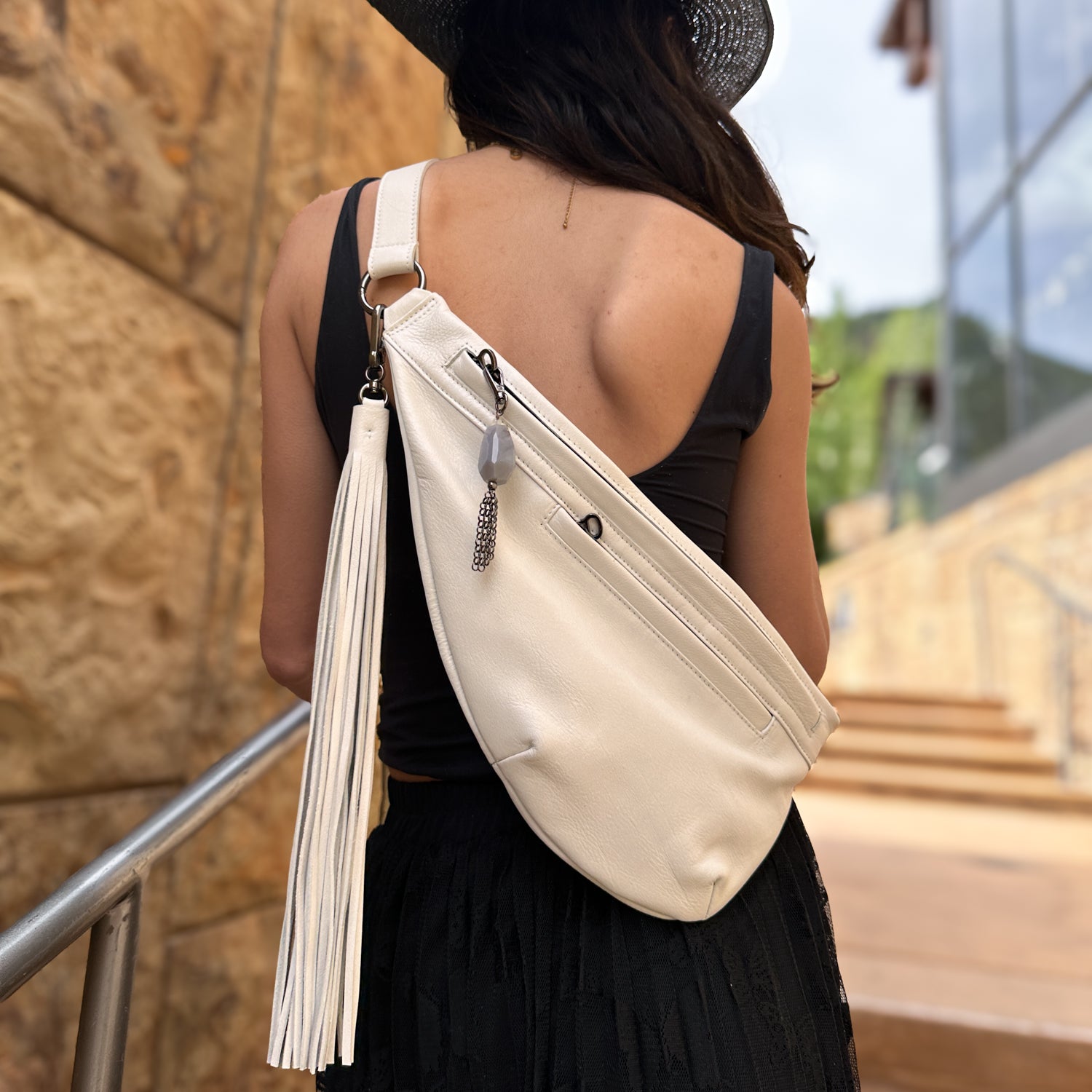 A person in a fashionable black outfit and hat is seen from behind, carrying the Sienna bag in pearl white with tassel details by evokewomen. They stand near a stone wall and railing, embodying effortless versatility.