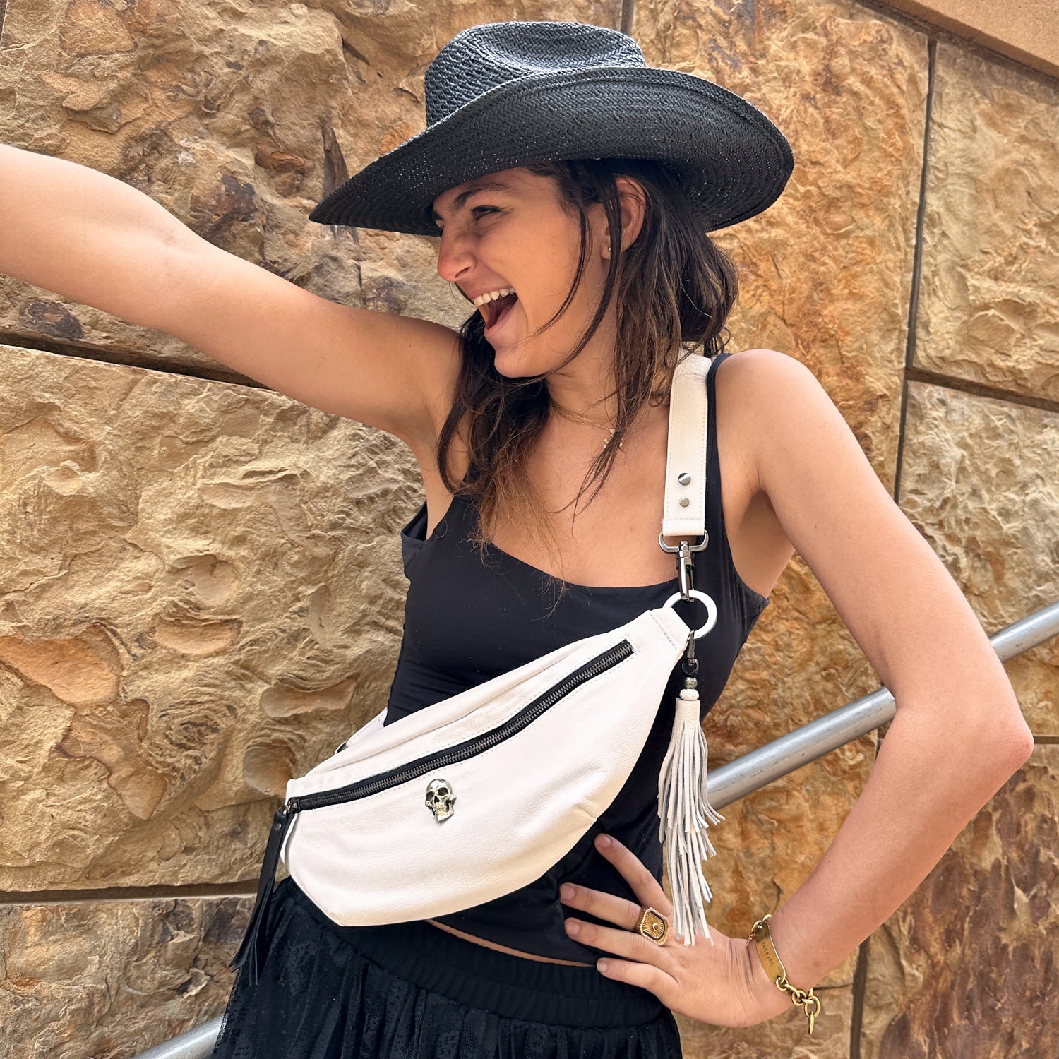 A person wearing a black hat and sleeveless top beams with delight, highlighting the STELLA - white - skull Italian leather sling bag from evokewomen, complete with a detachable tassel, set against a stone wall backdrop.