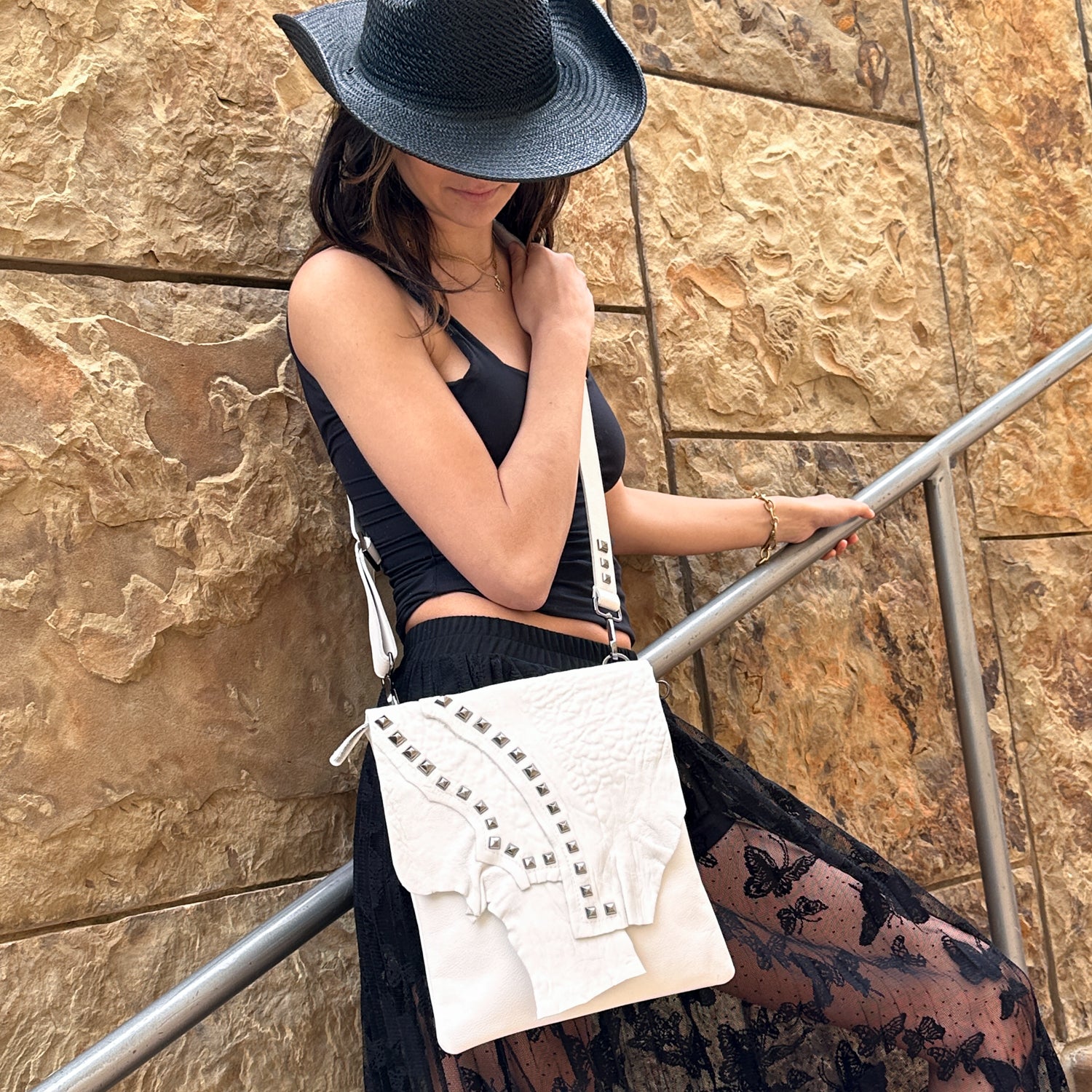 A person in a black hat and outfit leans on a railing, showcasing the elegant DRITA - white studded crossbody bag by evokewomen. The adjustable strap adds versatility to the look, while a stone wall in the background enhances the chic ensemble with timeless appeal.