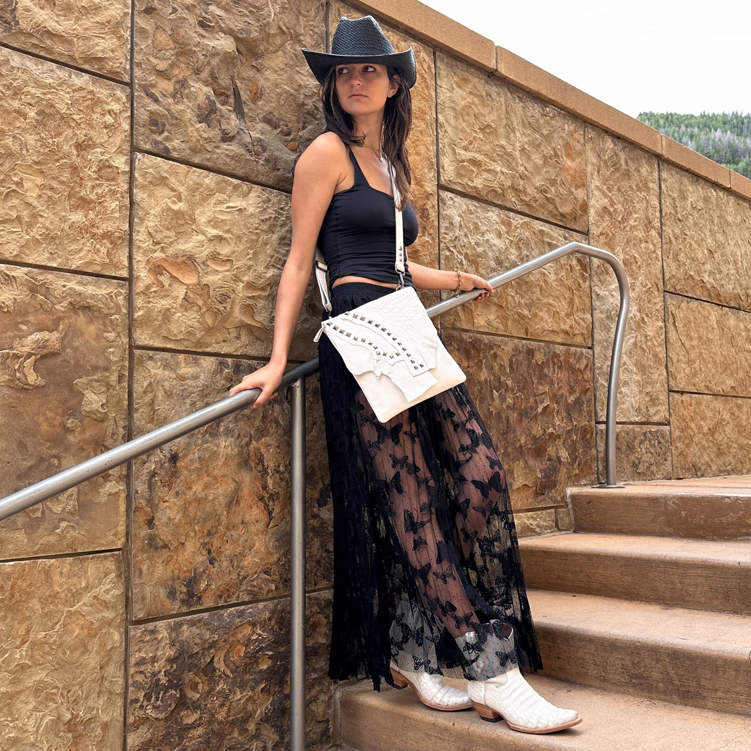 A person in a black hat, black top, lace skirt, white boots by the name of DRITA - white from evokewomen, and an Italian leather crossbody bag with an adjustable strap stands on stone steps by a textured wall.