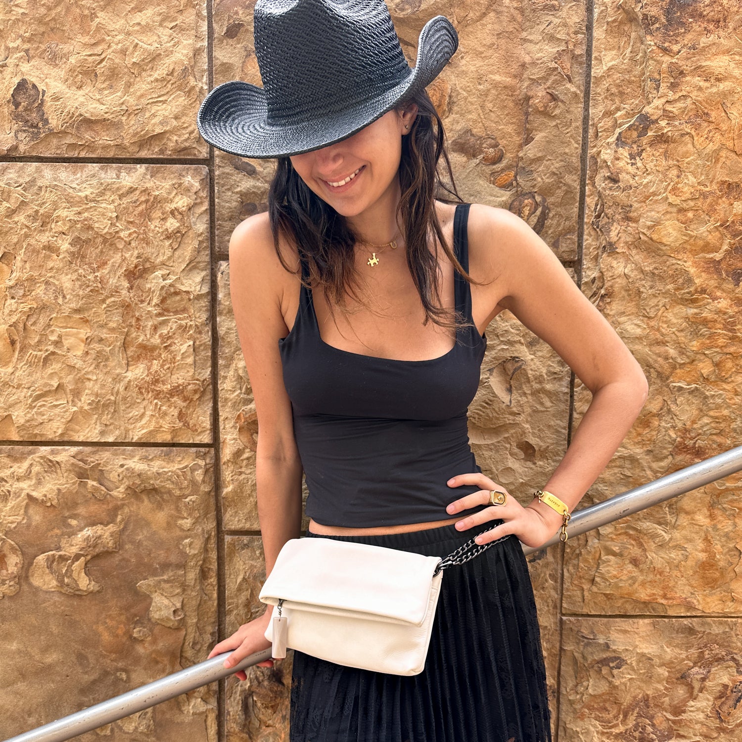 Wearing a sleeveless black top and hat, the person stands against a stone wall, smiling with the versatile Farah Fold-Over Clutch in pearl white by evokewomen across their waist.