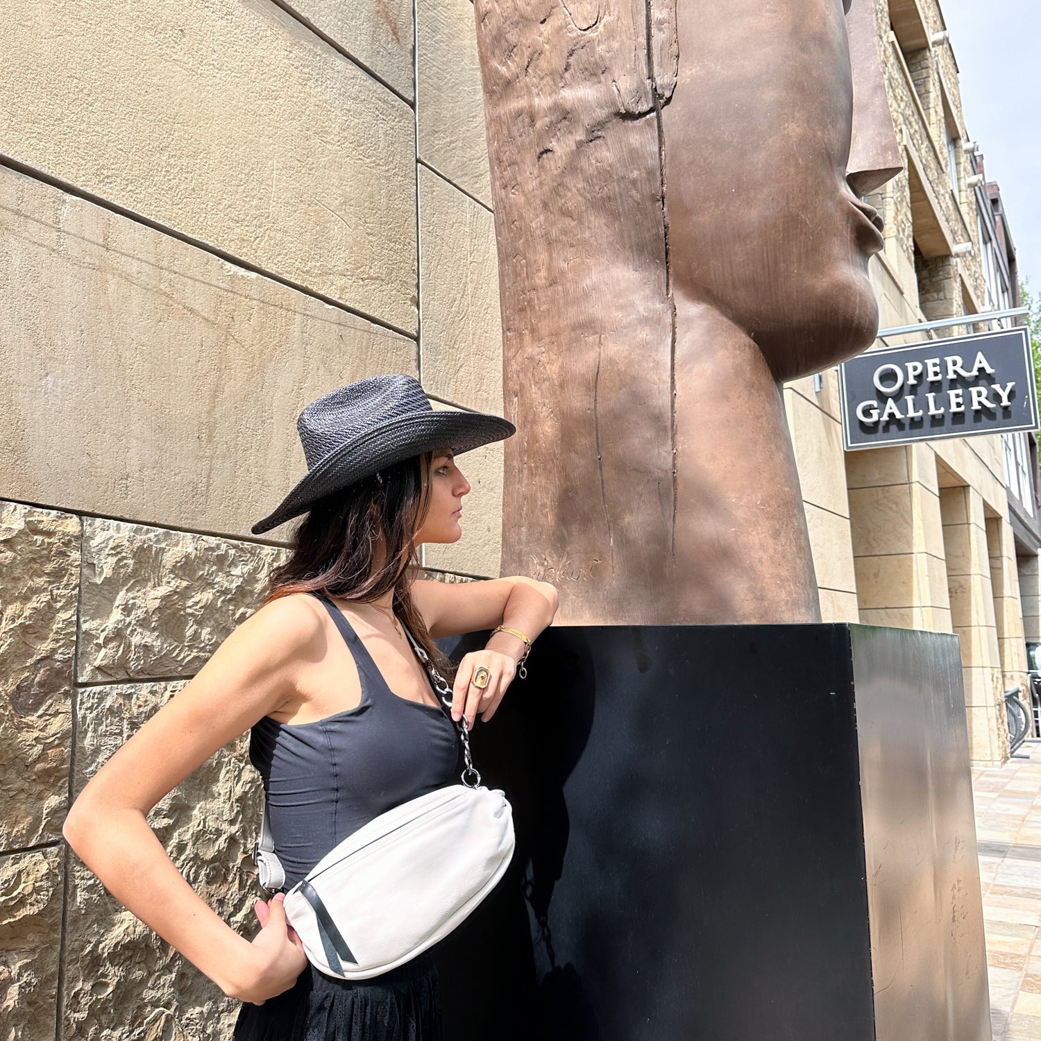 A woman in a black dress and hat stands next to a large sculpture head in front of Opera Gallery, exuding effortless chic with the LONDON - WHITE by evokewomen crossbody bag accentuating her style.