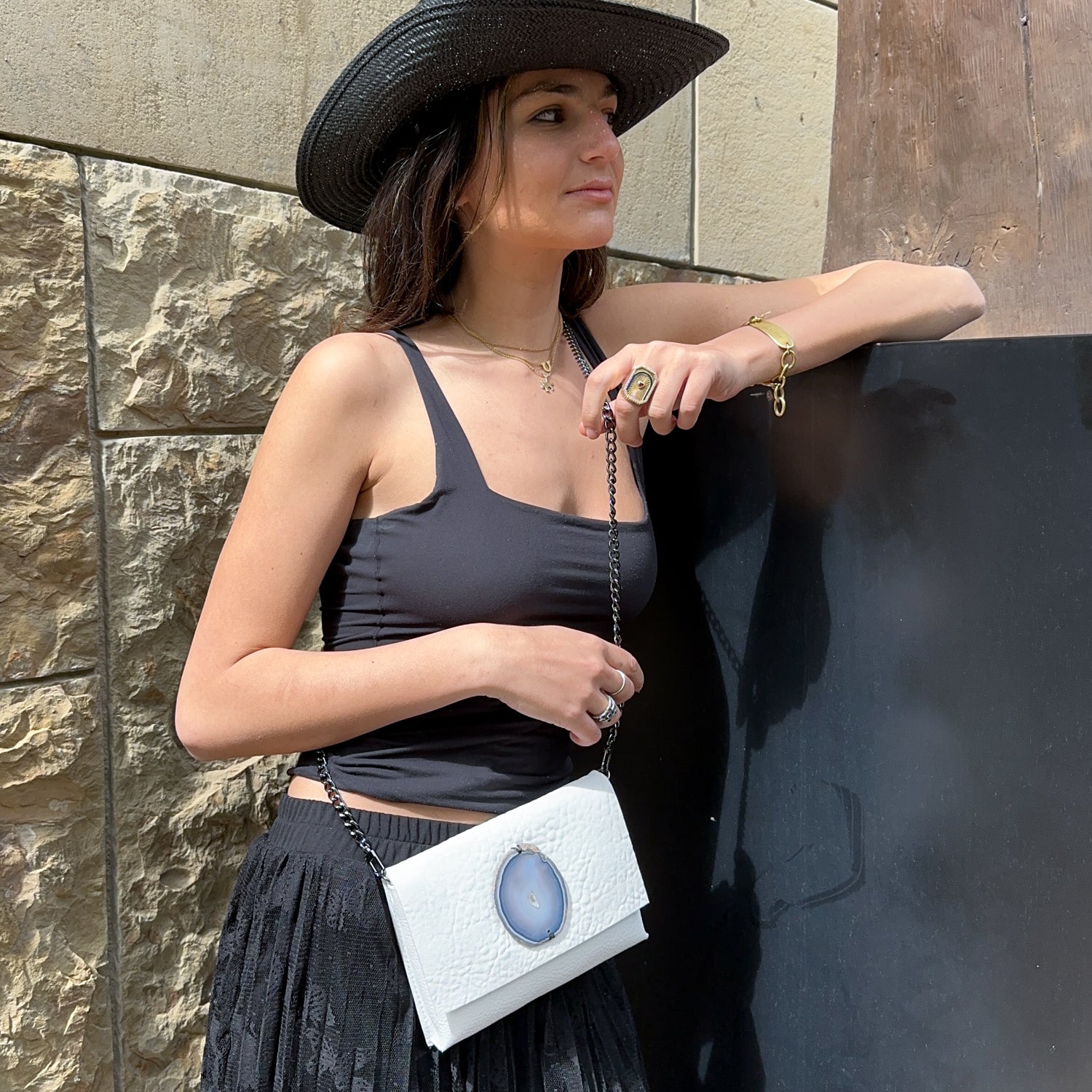 A woman dressed in a black hat, sleeveless top, and skirt leans casually against a stone wall, gracefully holding the BAHAMA - white handbag from evokewomen. The bag's versatile design enhances her outfit with a touch of elegance.