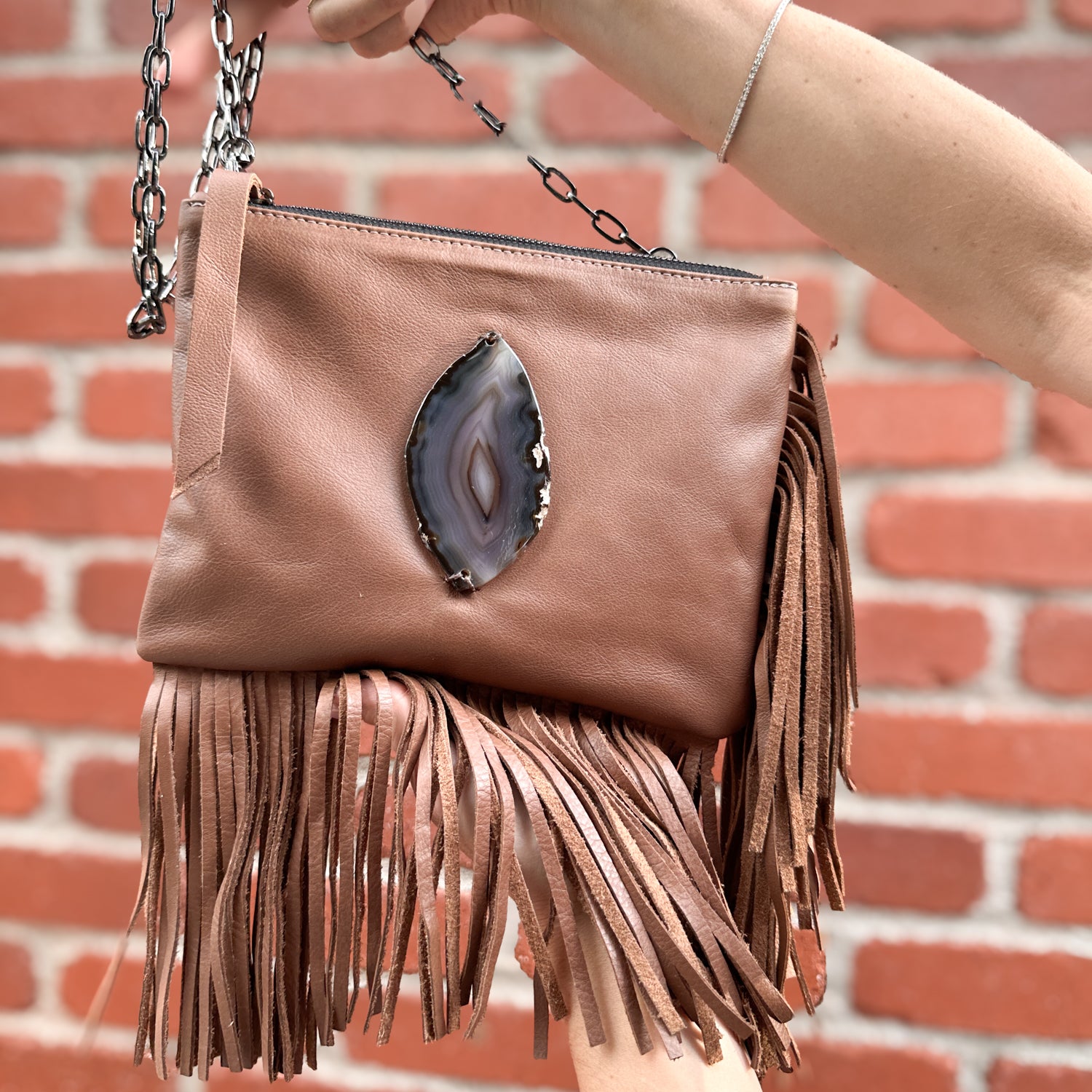 The Fringe Bag - Nutshell by evokewomen boasts brown leather fringes, a glittering gemstone accent, and a chic chain strap, perfectly showcased as it is elegantly held by a hand against a rustic brick wall.
