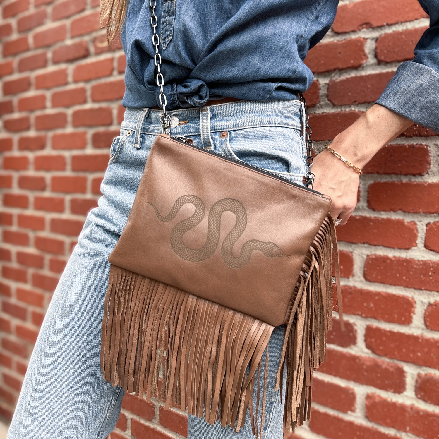 A close-up captures a person wearing a denim shirt and jeans, effortlessly holding the "Fringe bag - nutshell laser snake" by evokewomen, adorned with leather fringes and a snake design, set against a brick wall backdrop.