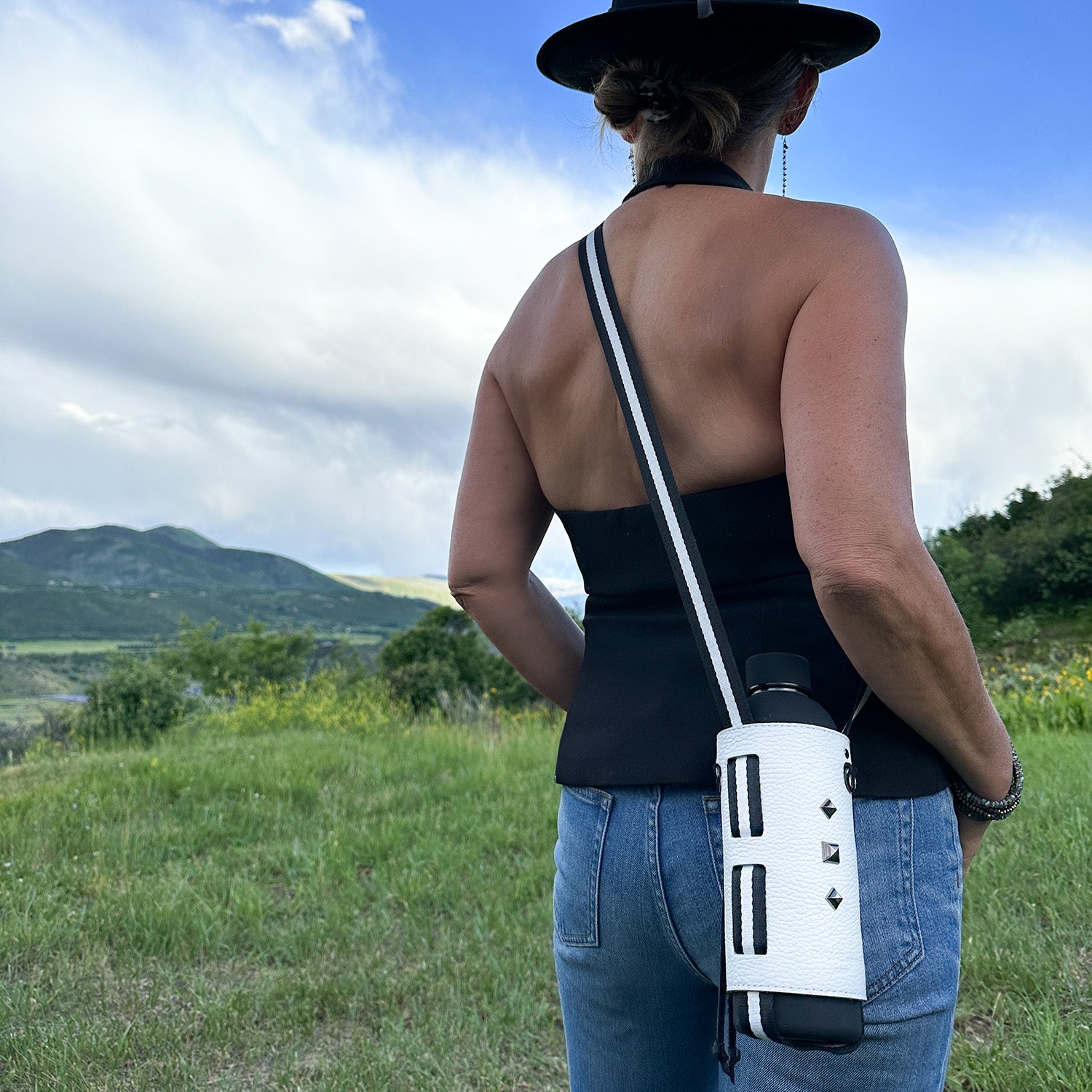An individual wearing a hat, black top, and jeans stands outdoors with the chic Bottle Carrier in white by evokewomen. The elegant carrier features an adjustable strap and complements the scenic backdrop of hills and clouds.