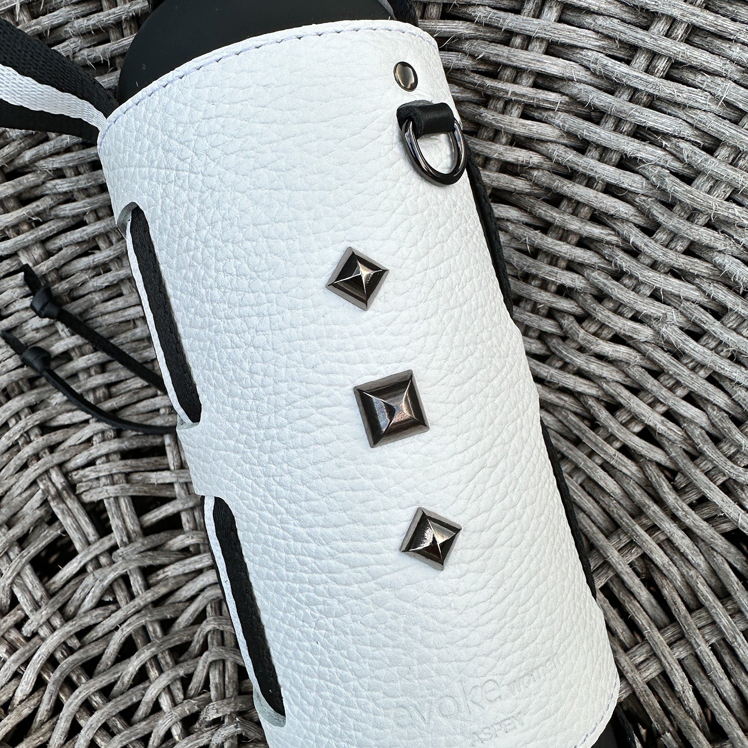 The Bottle Carrier - white by evokewomen is expertly crafted from white leather and showcases a textured cylindrical case adorned with metal studs and black accents. It is elegantly displayed on a woven surface and includes an adjustable shoulder strap for easy carrying.