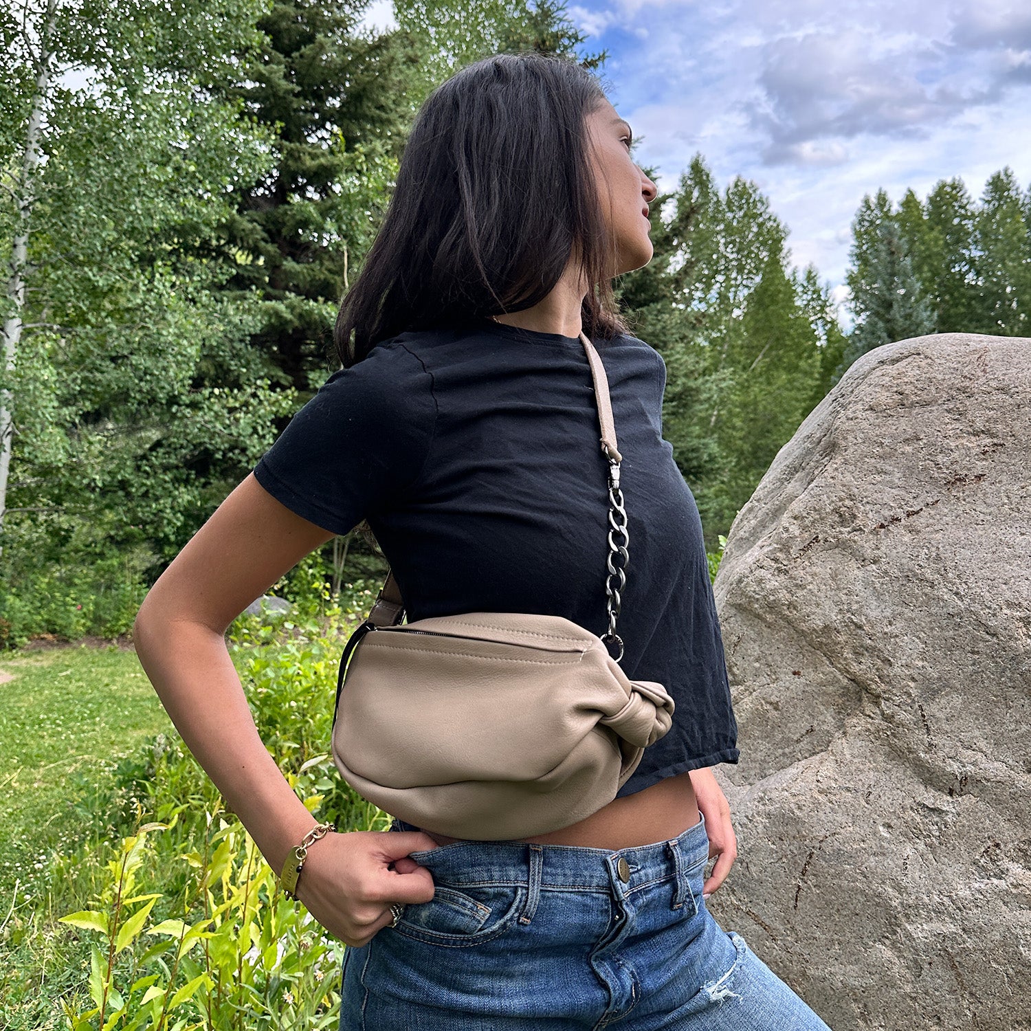 A fashion-forward individual in a black t-shirt and jeans stands outdoors near a large rock, carrying the evokewomen LONDON - SAND leather sling bag. Trees and greenery provide a lush backdrop.